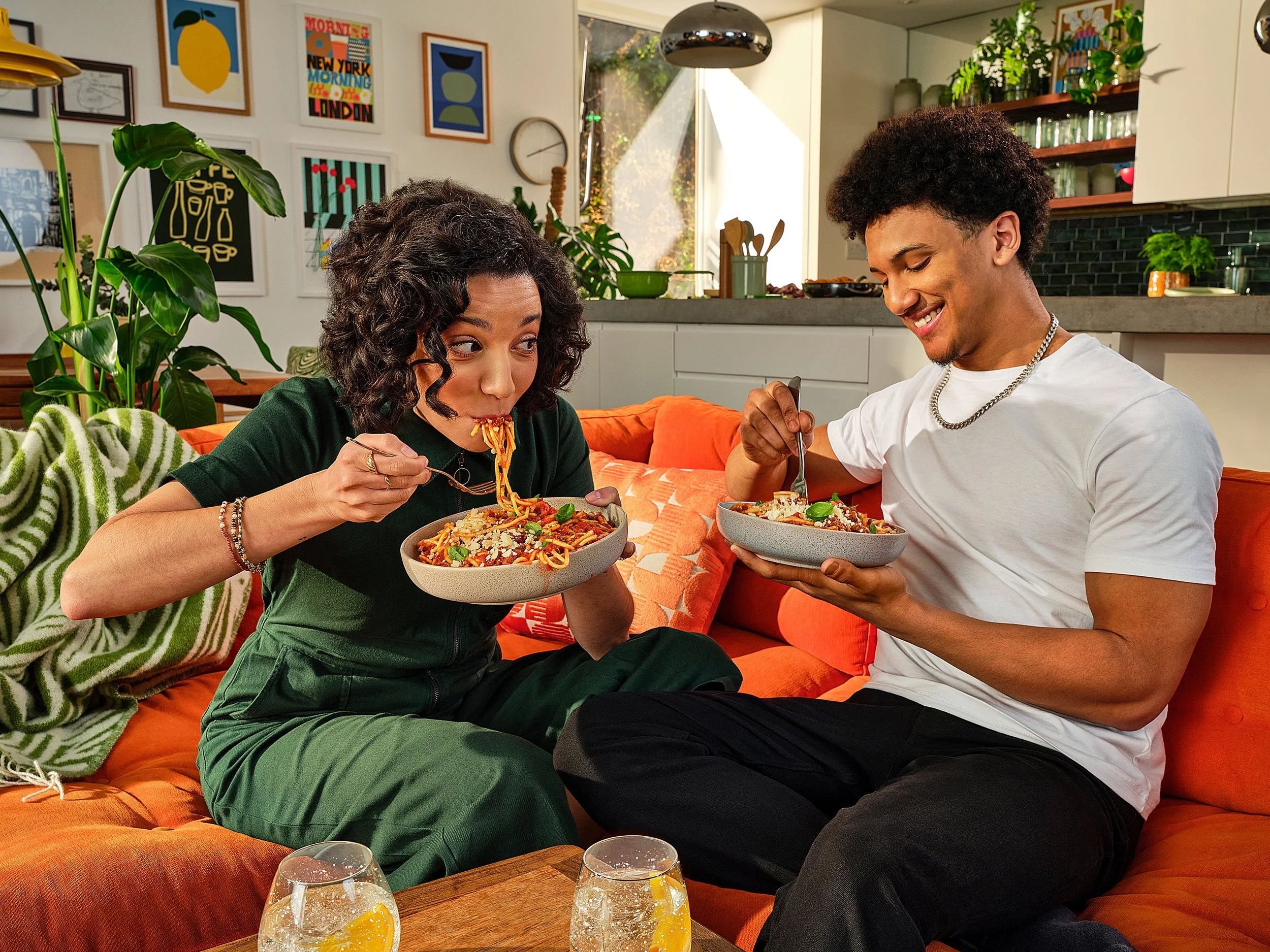 A young woman and a young man are sitting on an orange couch in a modern living room, eating spaghetti. The woman has curly dark hair, and the man has a large curly afro. They are smiling and appear to enjoy their meal. There are several glasses of w