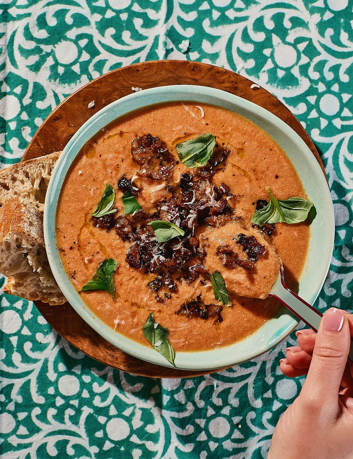 A bowl of tomato soup garnished with basil leaves and crispy bacon bits, served with a slice of rustic bread on a wooden plate, on a patterned teal and white tablecloth.