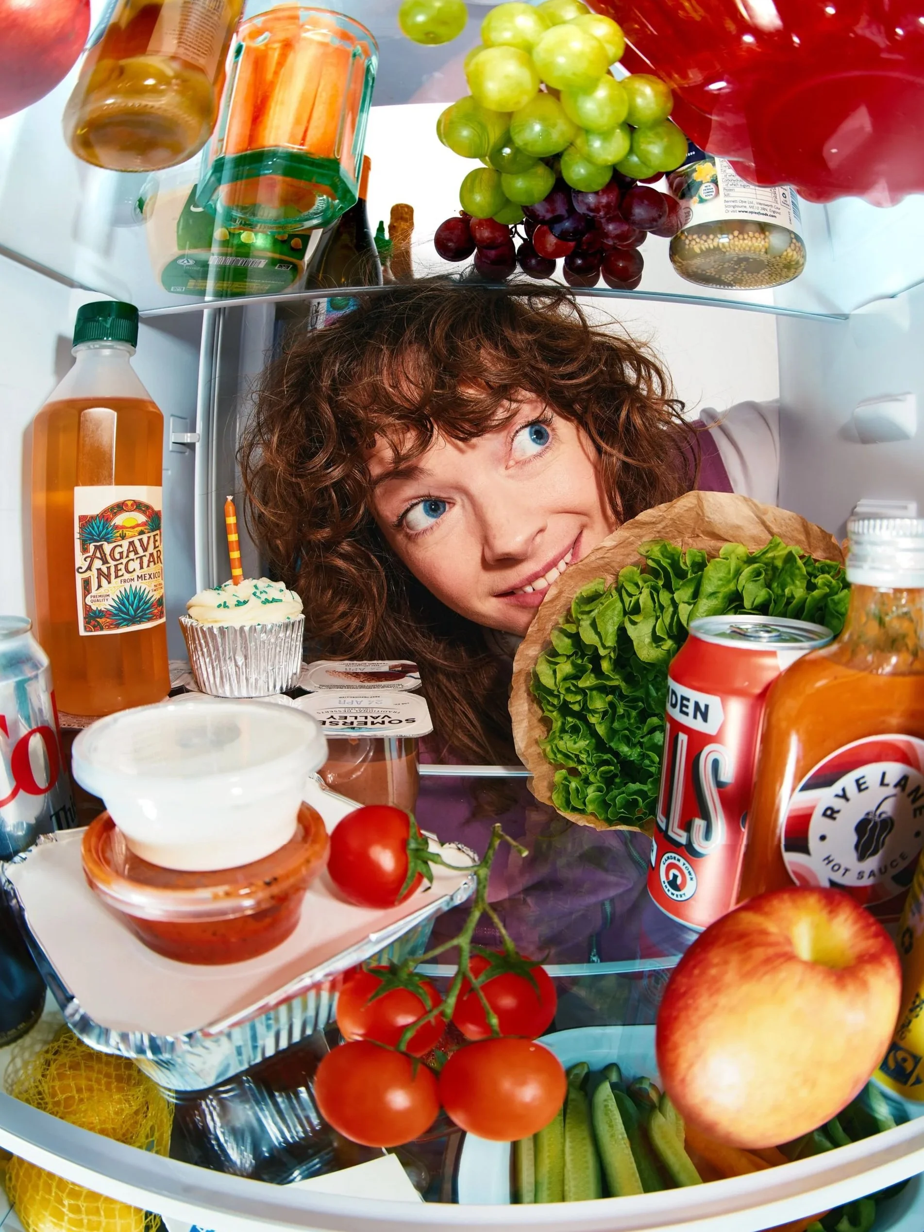 A portrait of photographer Jo Sidey, peering inside a refrigerator door filled with various food items including, agave, a cupcake with a candle, tomatoes, apples, lettuce, cucumber slices, grape juice, and other condiments and food containers.