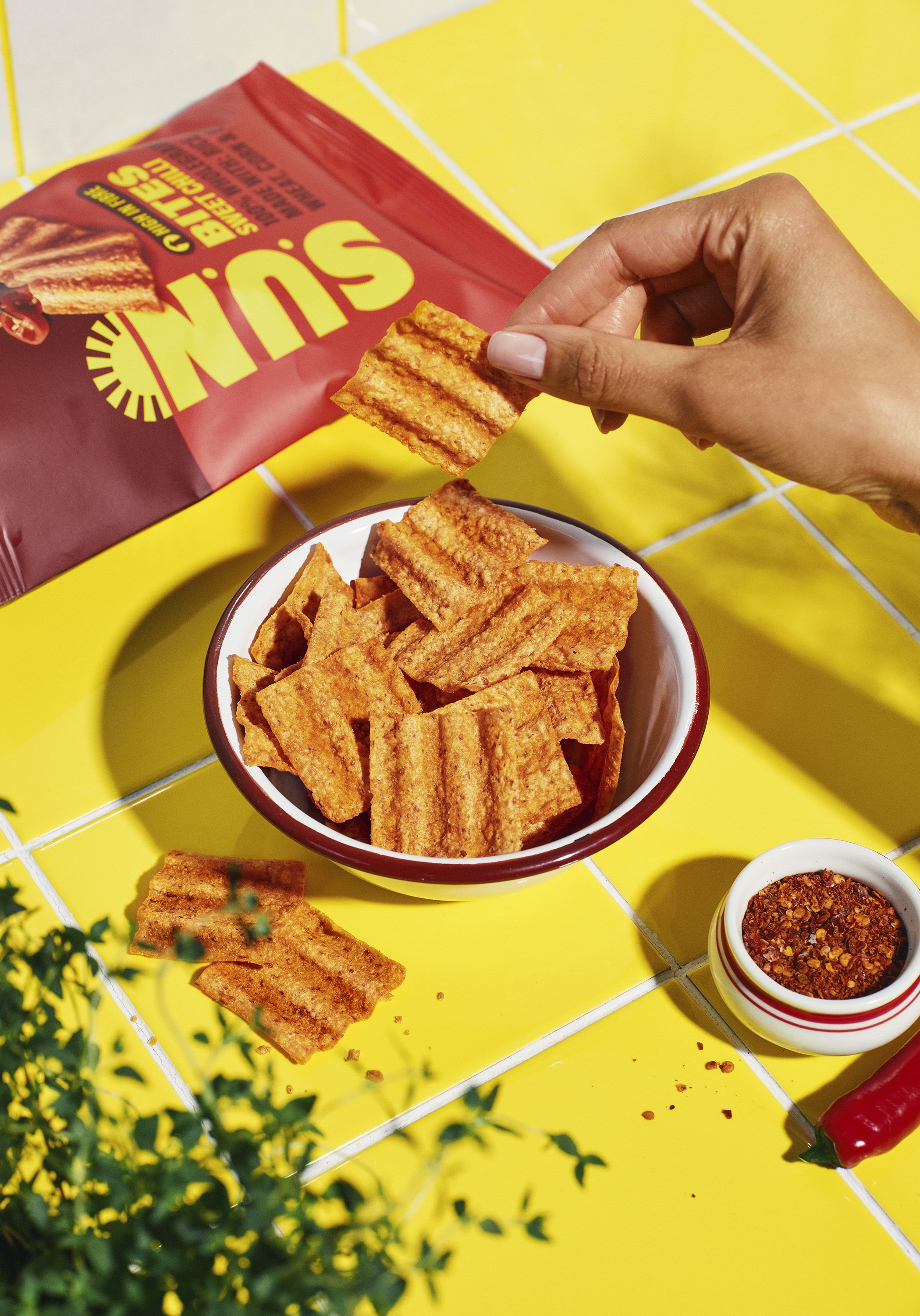 Basket of crispy pork cracklings on a yellow tiled table with a small bowl of red chili flakes, a red hot chili, and a bag of pork cracklings in the background.