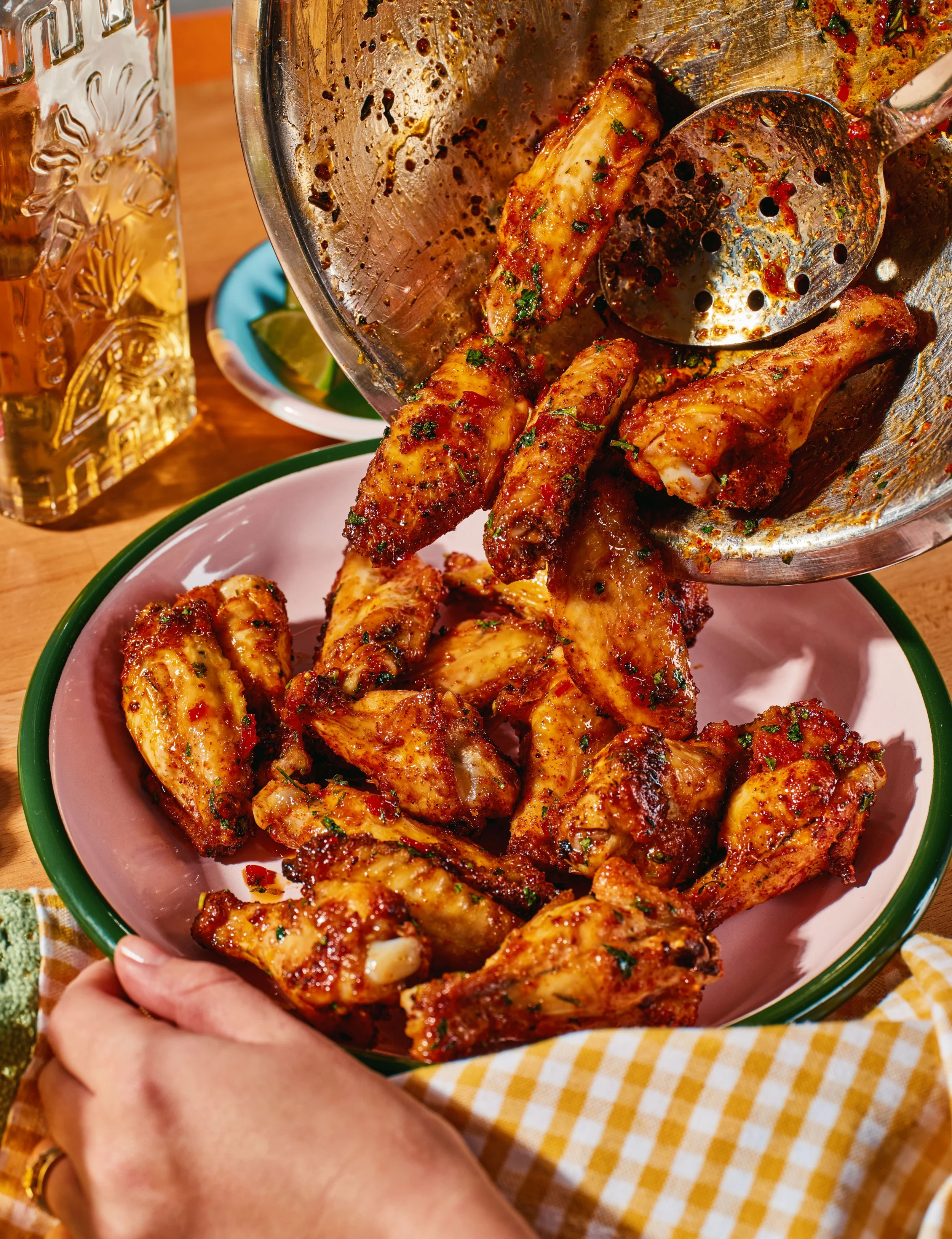 Close-up of cooked and seasoned chicken drumsticks in a pink bowl with a green rim, being held by a person's hand. In the background, a glass of beer, a small dish with lime wedges, and a yellow and white checkered cloth are visible.