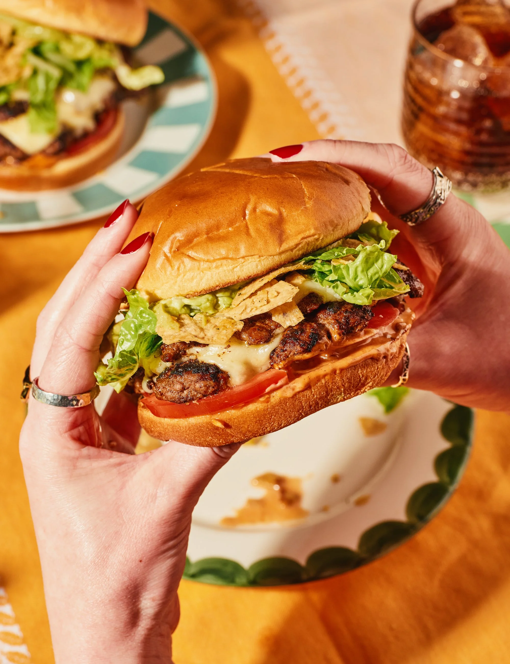 Person holding a cheeseburger with lettuce, tomato, and grilled beef patty, with a plate of another burger and a glass of soda in the background.