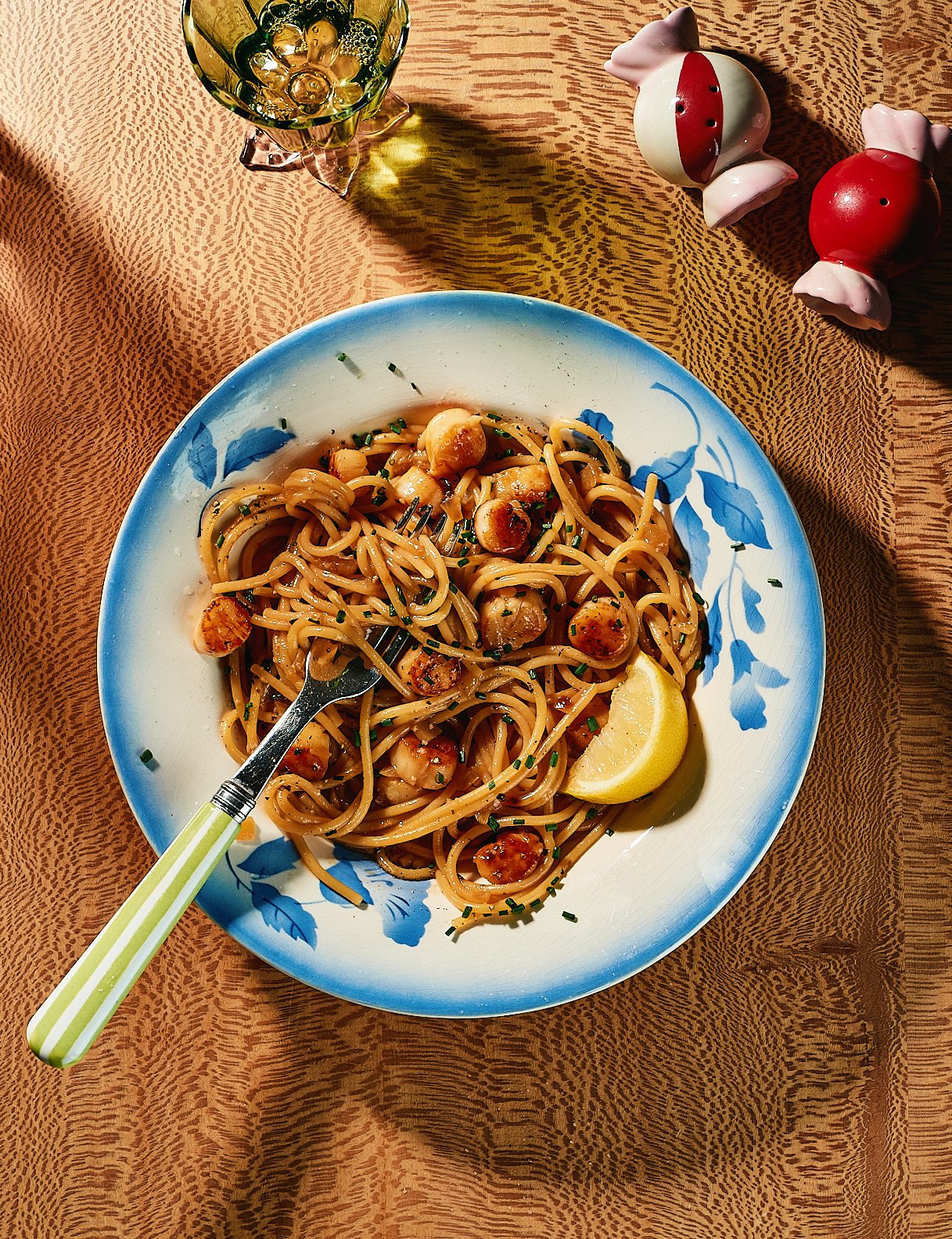 A plate of pasta with scallops, a lemon wedge, and chopped herbs on a blue and white dish, placed on a patterned orange tablecloth. A fork with a green and white handle is in the pasta. In the background, there is a glass of white wine and two cerami