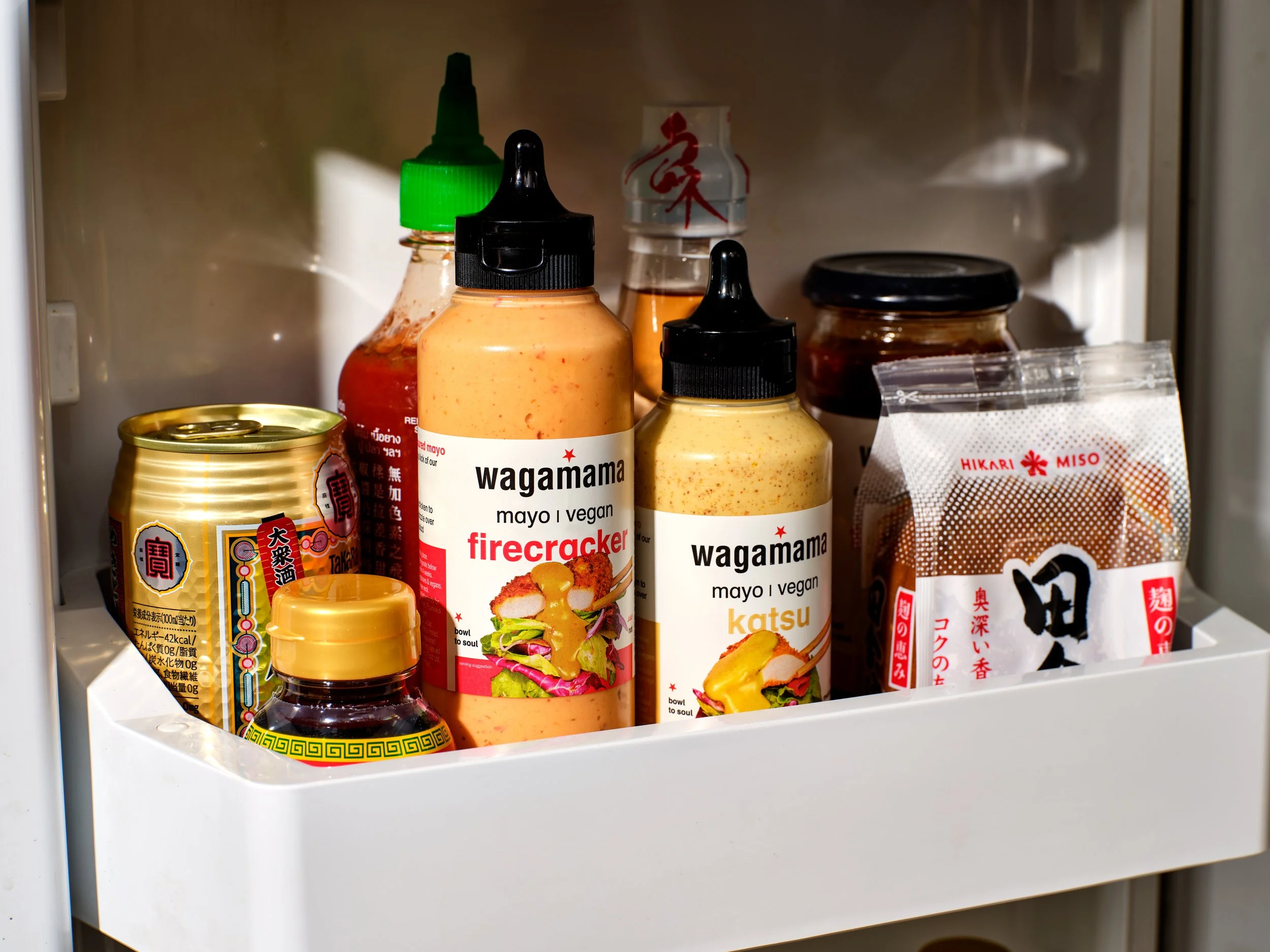 A close-up view of asian condiments and sauces inside a refrigerator drawer, including bottles of Wagamama vegan mayo, a can, and a package of miso paste.