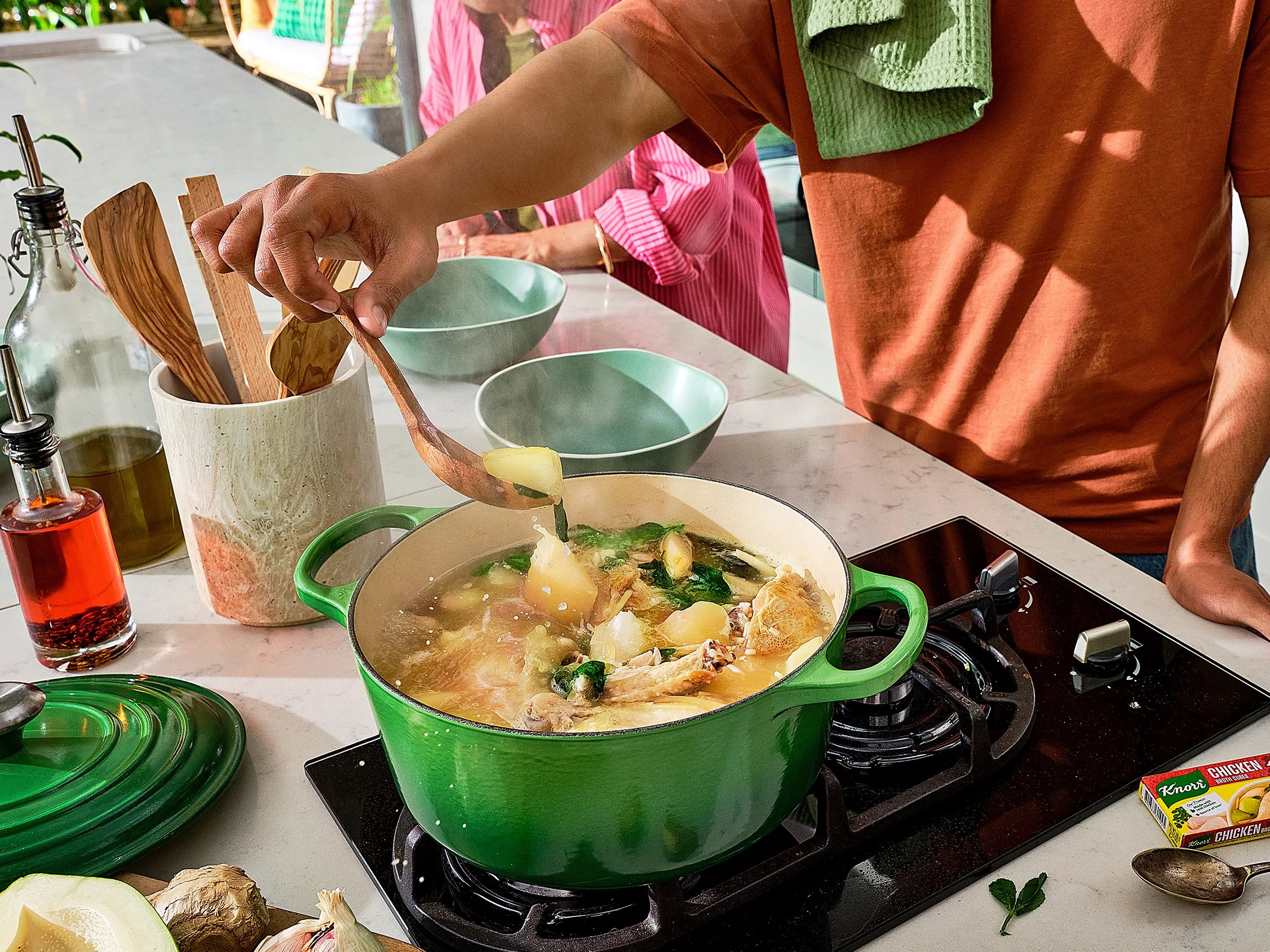 A person is adding ingredients into a green pot on the stove, with a soup or stew inside. The kitchen counter has various cooking items and vegetables, with two empty green bowls nearby.
