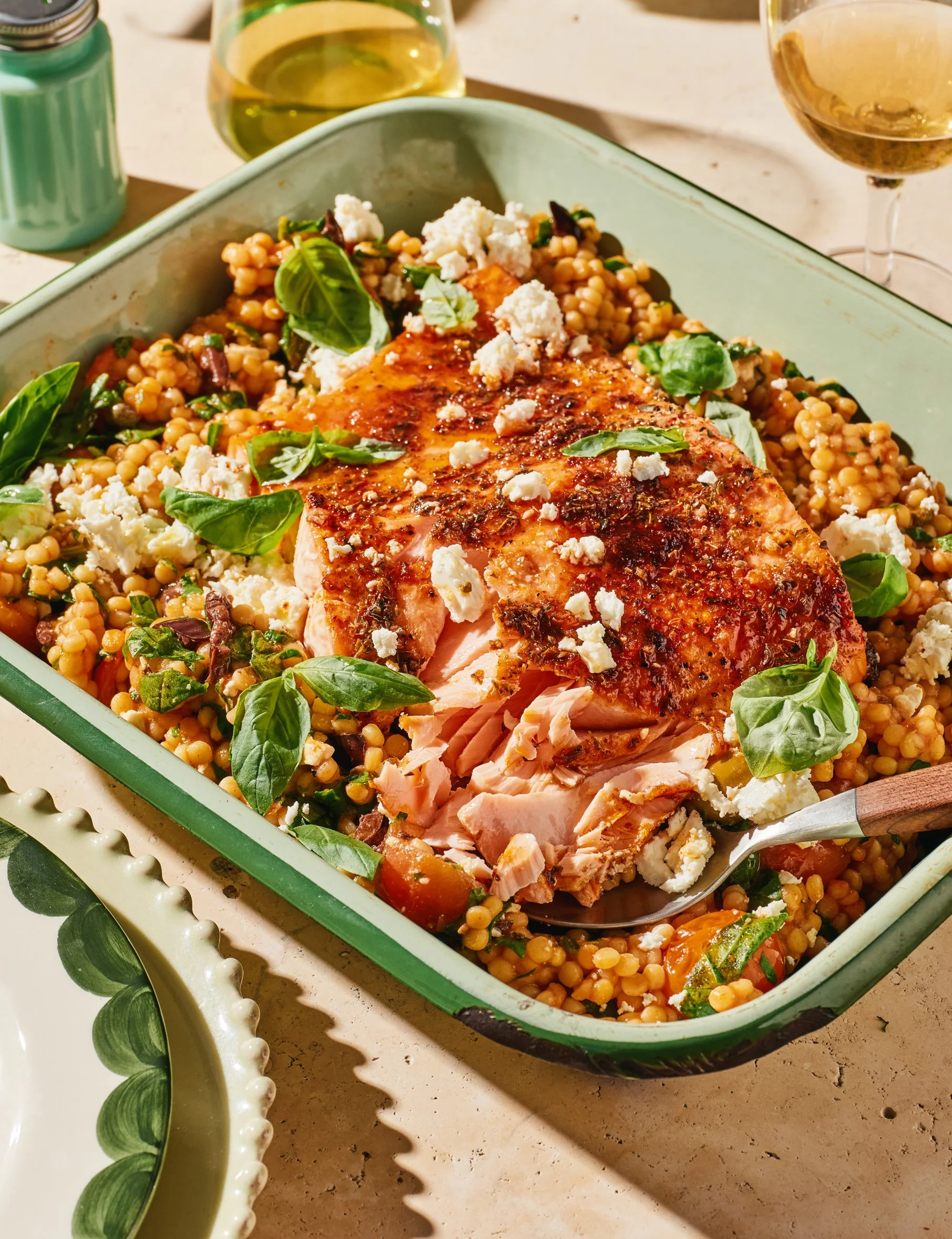 A baked salmon fillet topped with herbs and seasoning, served on a bed of cooked couscous, garnished with fresh basil and crumbled cheese, in a green ceramic dish. Two glasses of white wine and a salt shaker are visible in the background.