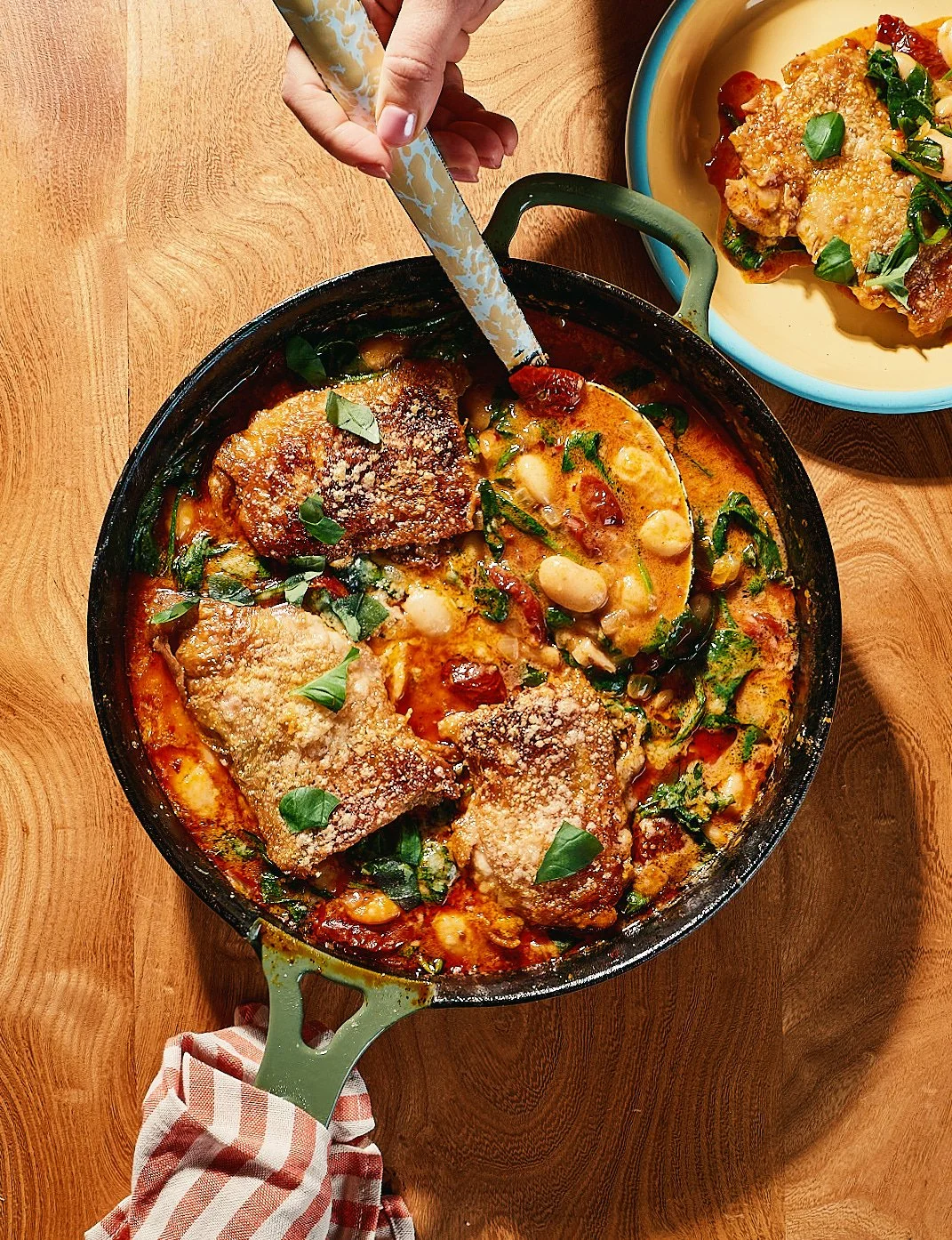 A pot of baked pasta with chicken, cheese, and vegetables, garnished with basil, with a hand holding a spoon and a plate of more baked pasta on a wooden table.