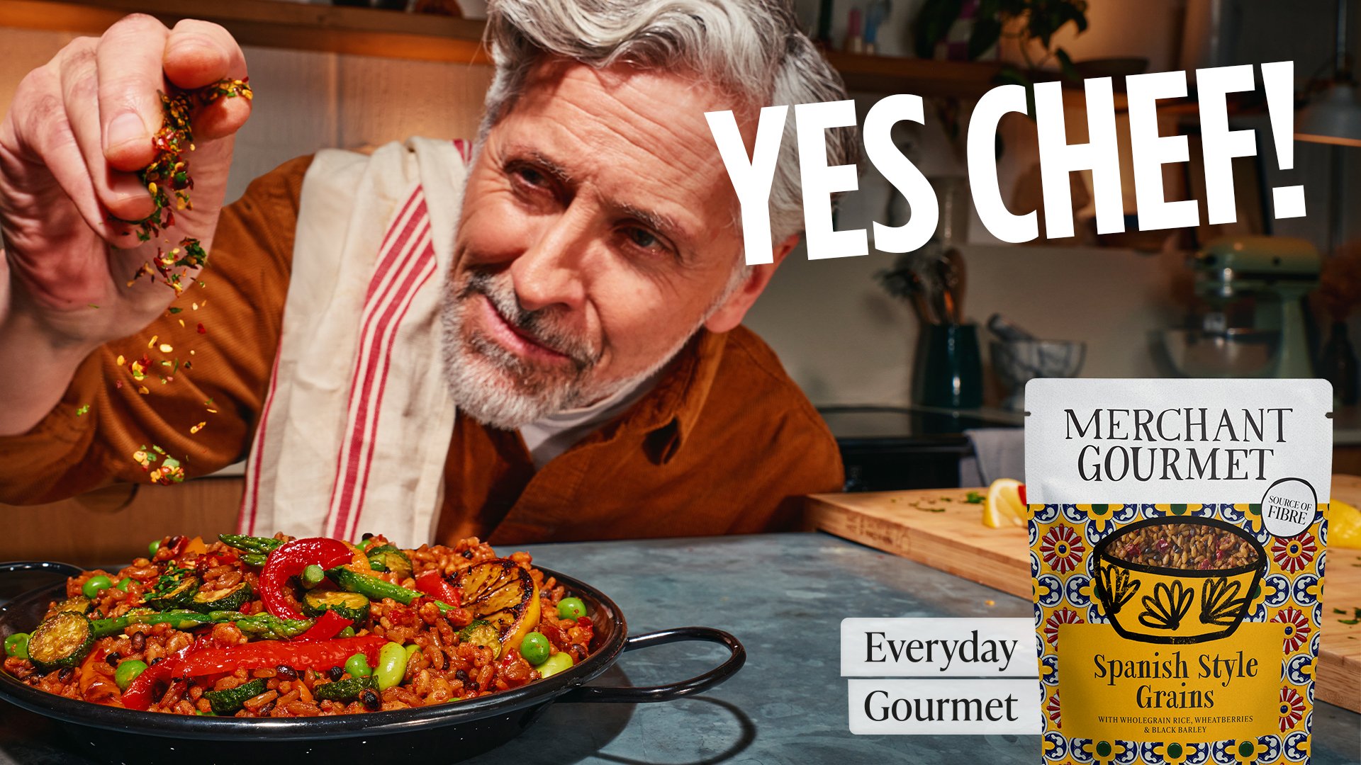 A man with gray hair and beard, wearing an orange shirt, is sprinkling colorful seasoning onto a plate of Spanish style grains with vegetables. There is a package of grain mix labeled "Spanish Style Grains" on the counter nearby, and a cutting board,