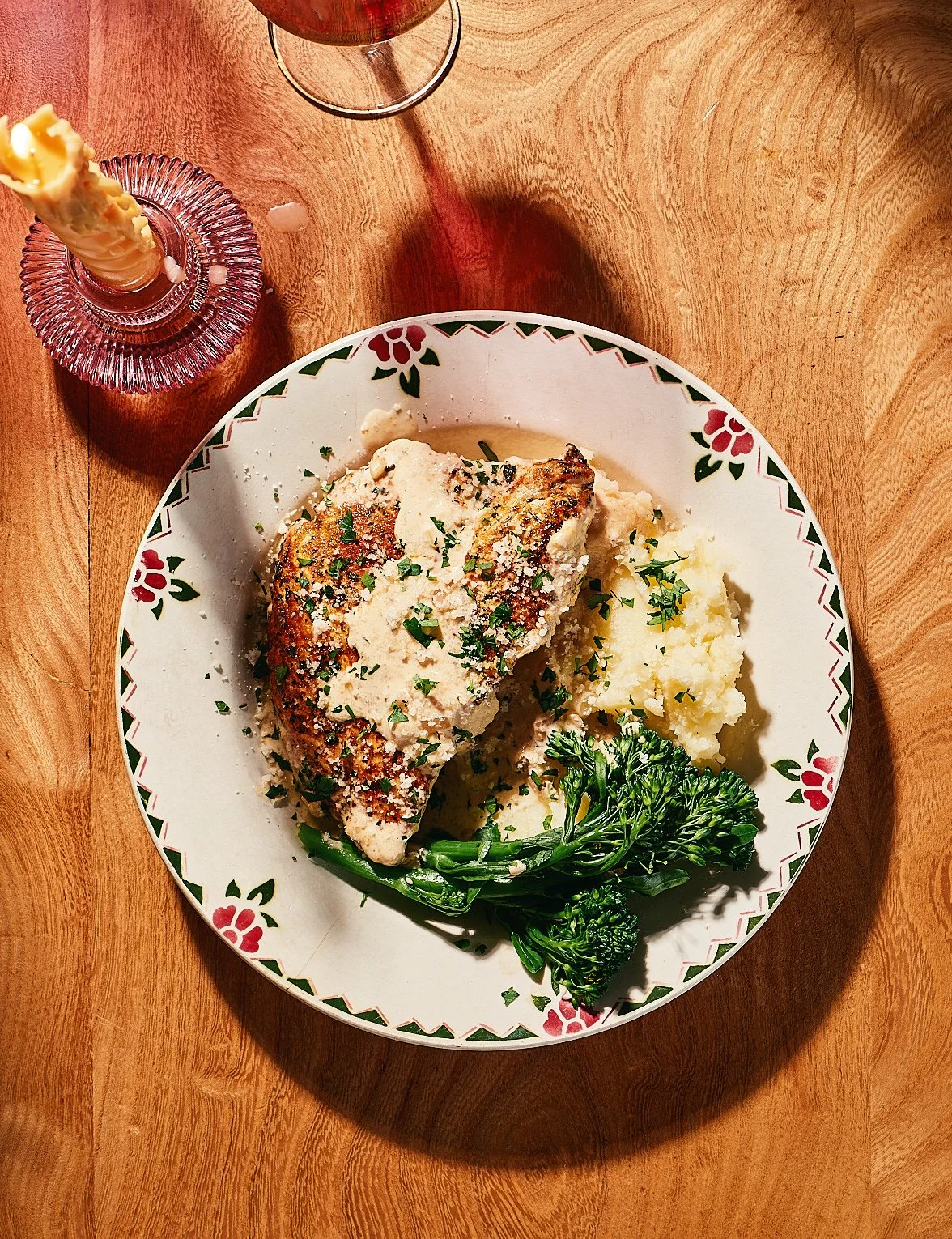 A plate of cooked chicken topped with gravy, mashed potatoes, and steamed broccoli. There is a glass of red wine and a candle on the wooden table.