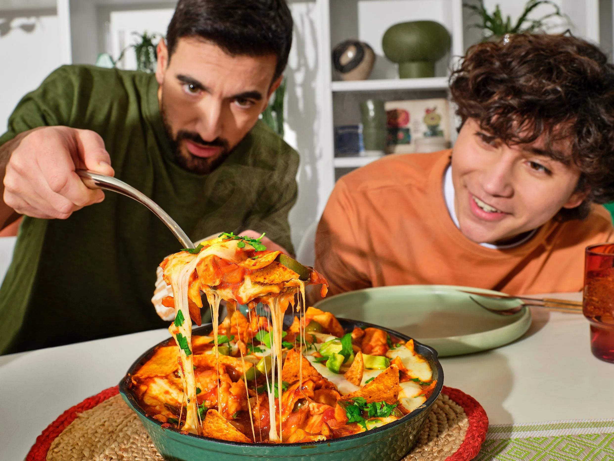 A man and a boy are sitting at a table with a large bowl of nachos topped with cheese, chili, and vegetables, sharing a meal.