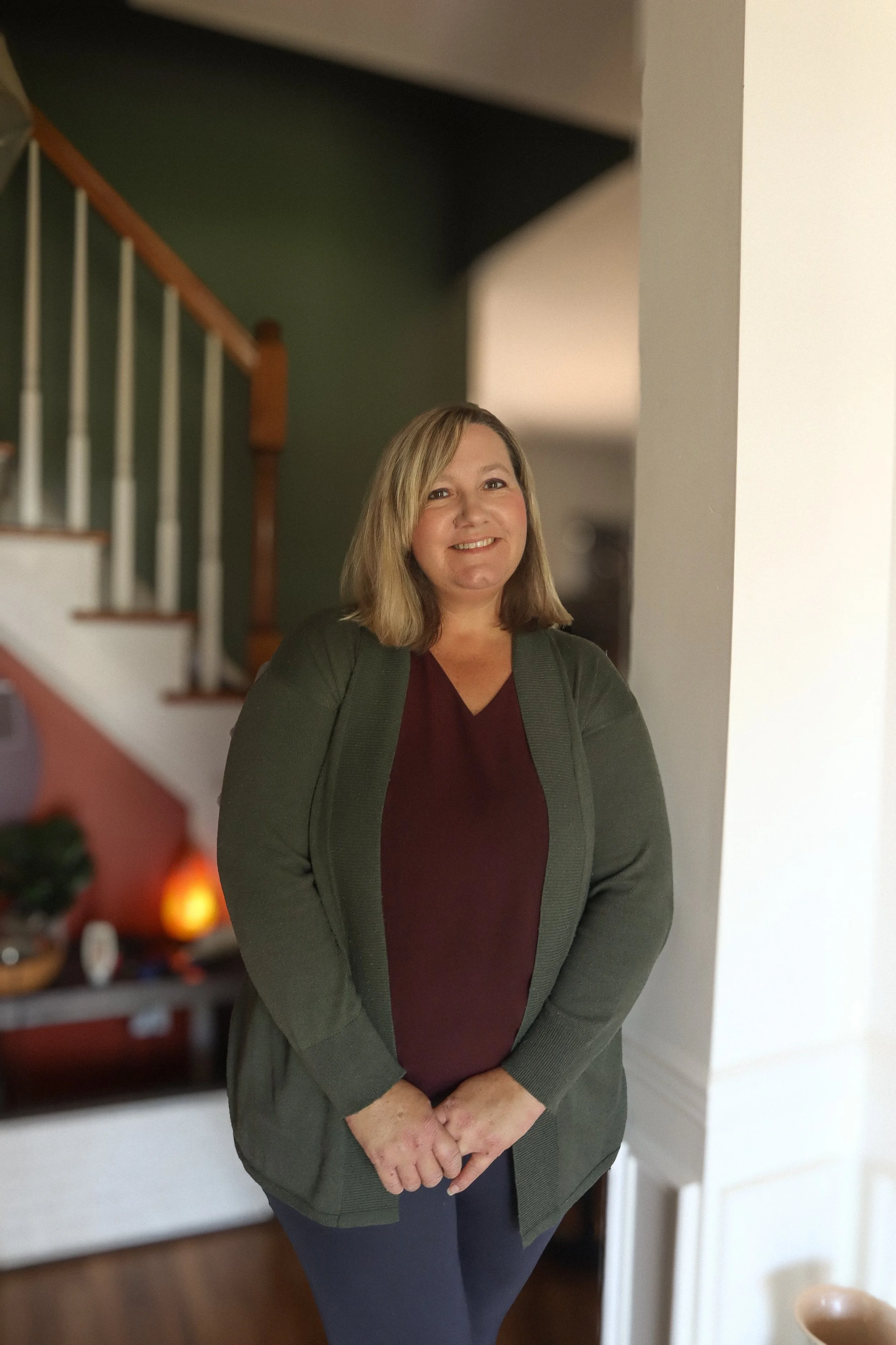 A woman with shoulder-length blonde hair standing indoors, smiling, wearing a dark red top and a green jacket.