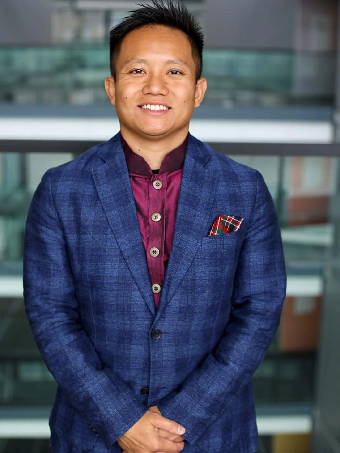 Photo of Danny An Khoi Vu, a smiling man wearing a blue lazer and a burgundy shirt, standing with hands clasped in front of him.