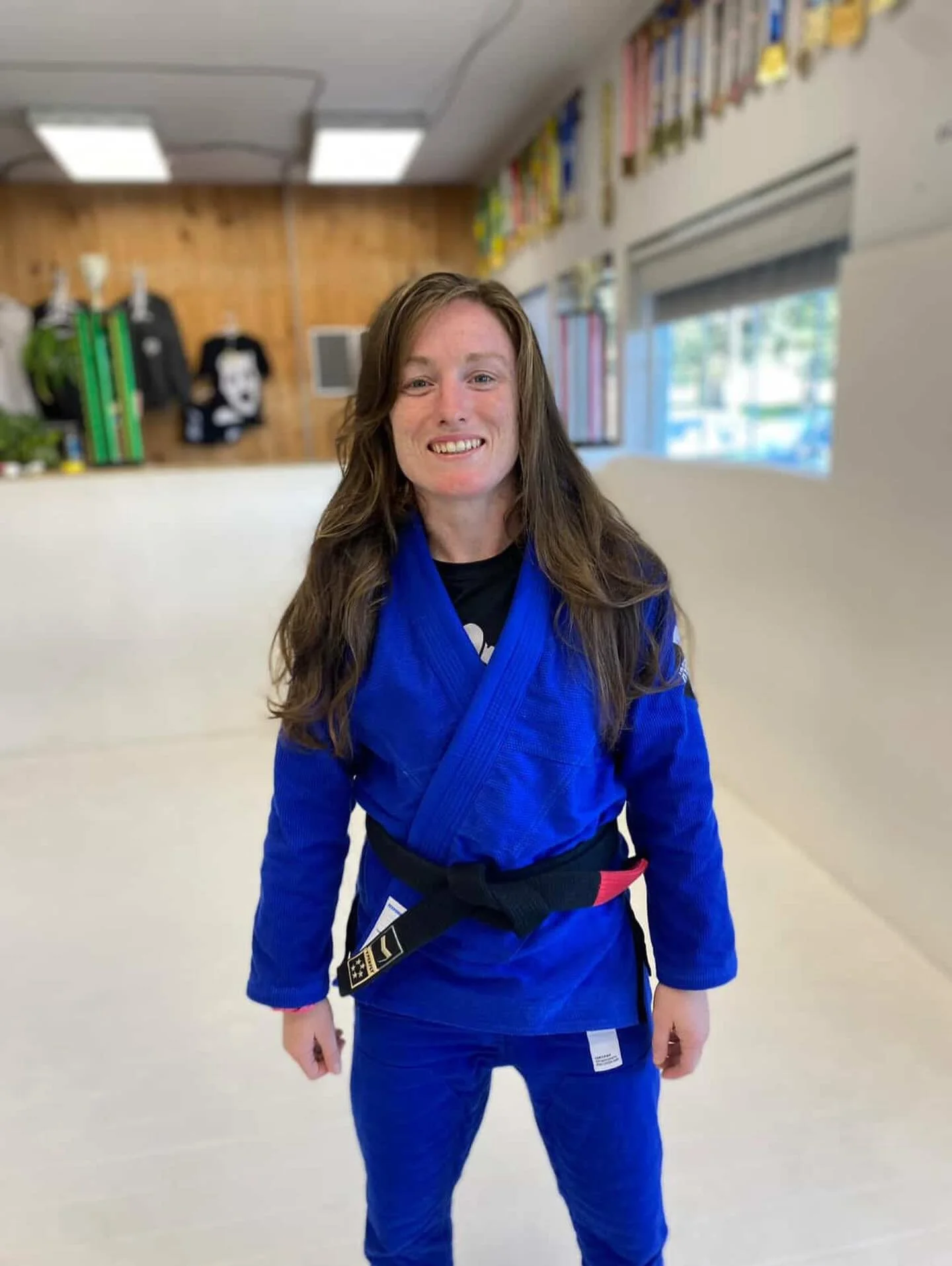 Woman wearing a blue jiu-jitsu gi with a black belt, standing and smiling inside a martial arts training gym.
