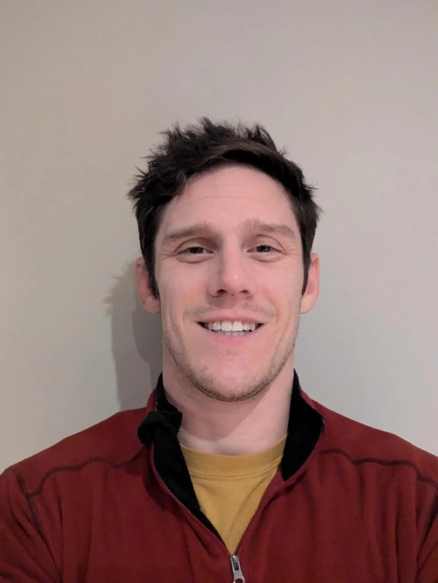 Portrait of a smiling man with short dark hair wearing a red sweater over a yellow shirt, standing against a plain background 