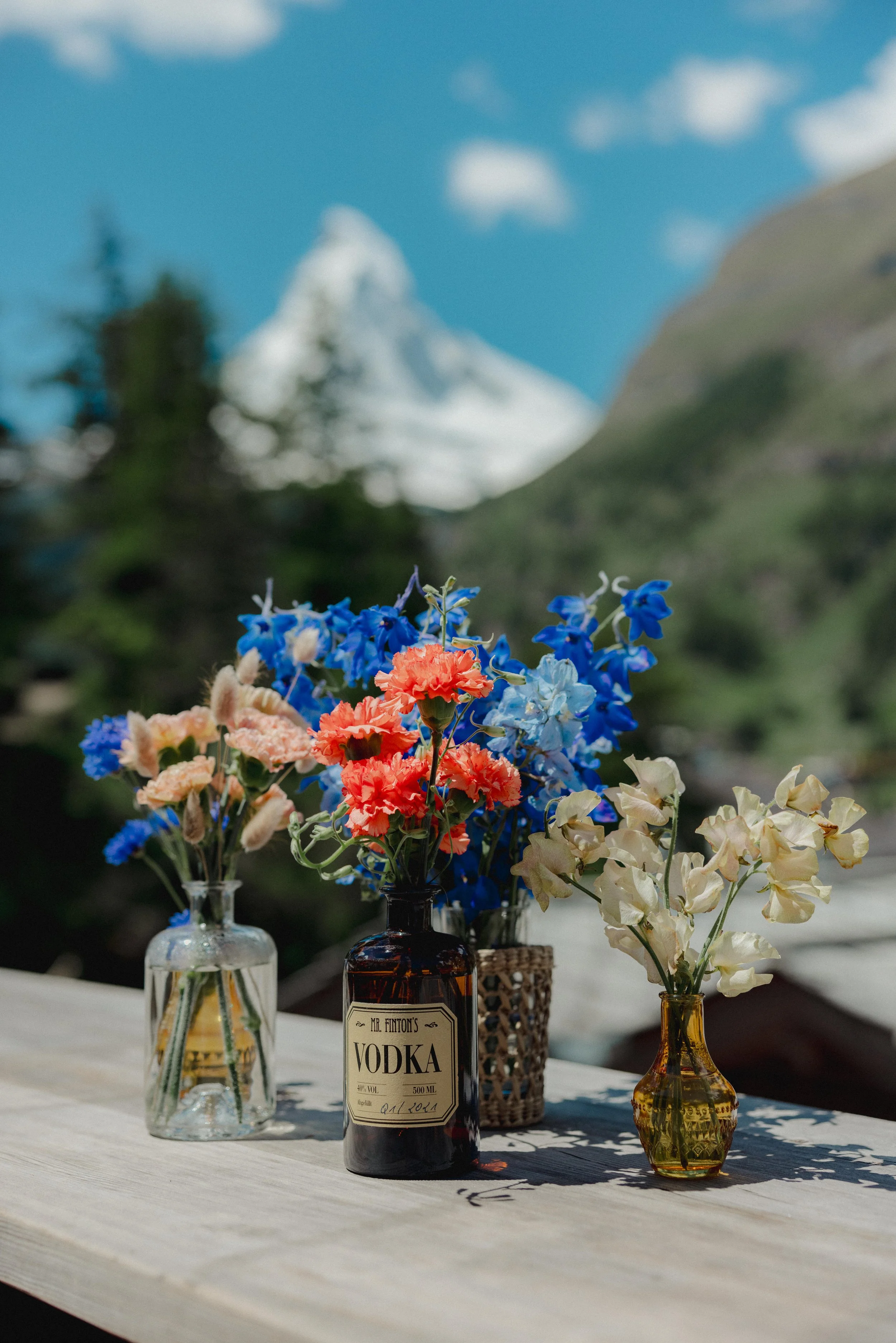 Colorful flower arrangements in decorative bottles on wooden table outdoors with mountain background for a mountain wedding.