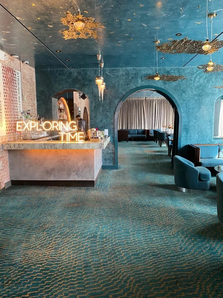 Interior of a stylish lounge with blue decor, ornate lighting, and a bar area. "Exploring Time" neon sign is visible. The ceiling has decorative gold elements. Arched doorways lead to seating areas with curtains. VIP press and media event for luxury 