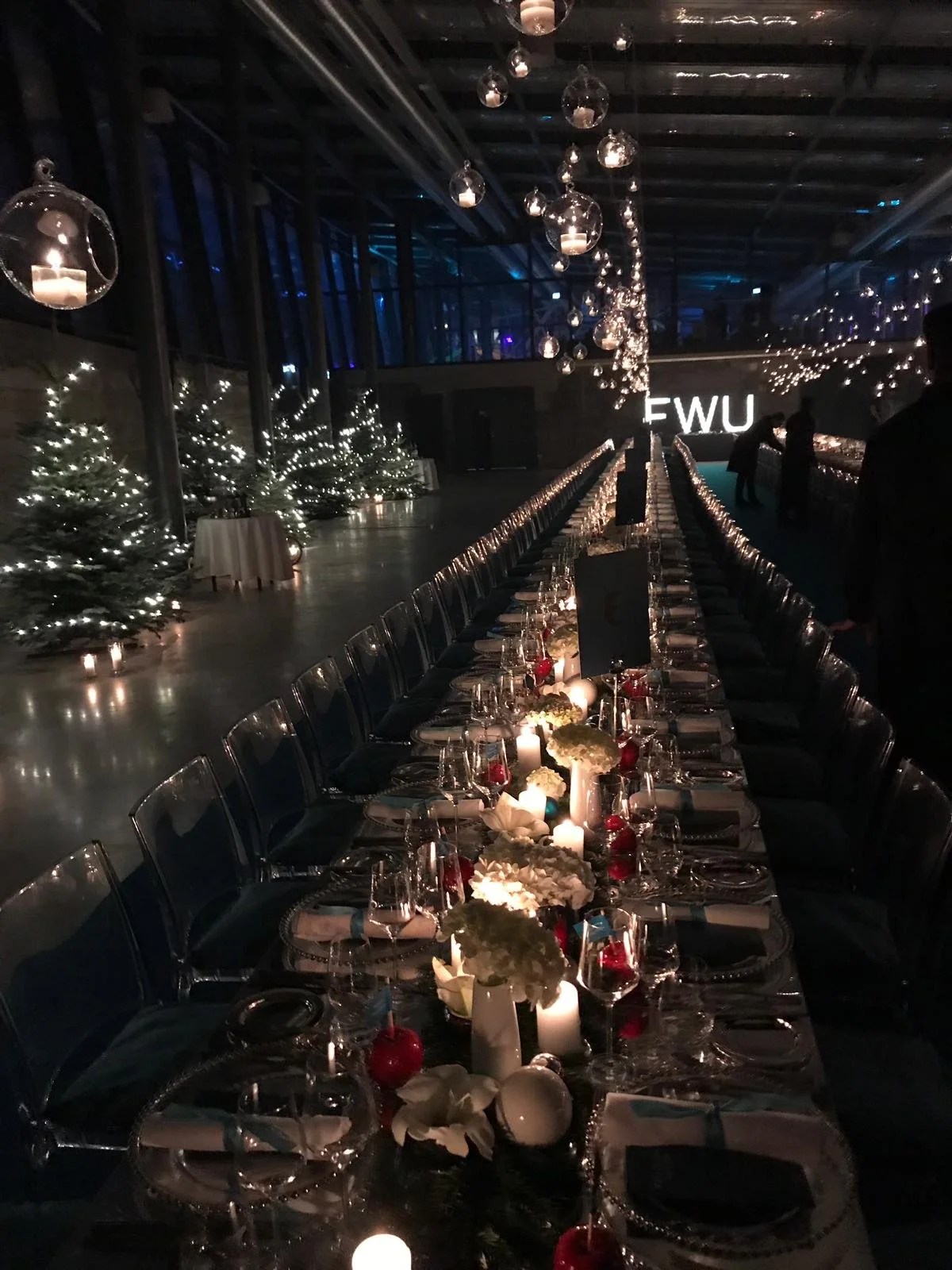 Long elegantly decorated dining table with candles, glassware, and floral centerpieces in a dimly lit venue with festive string lights and Christmas trees during a Christmas party.