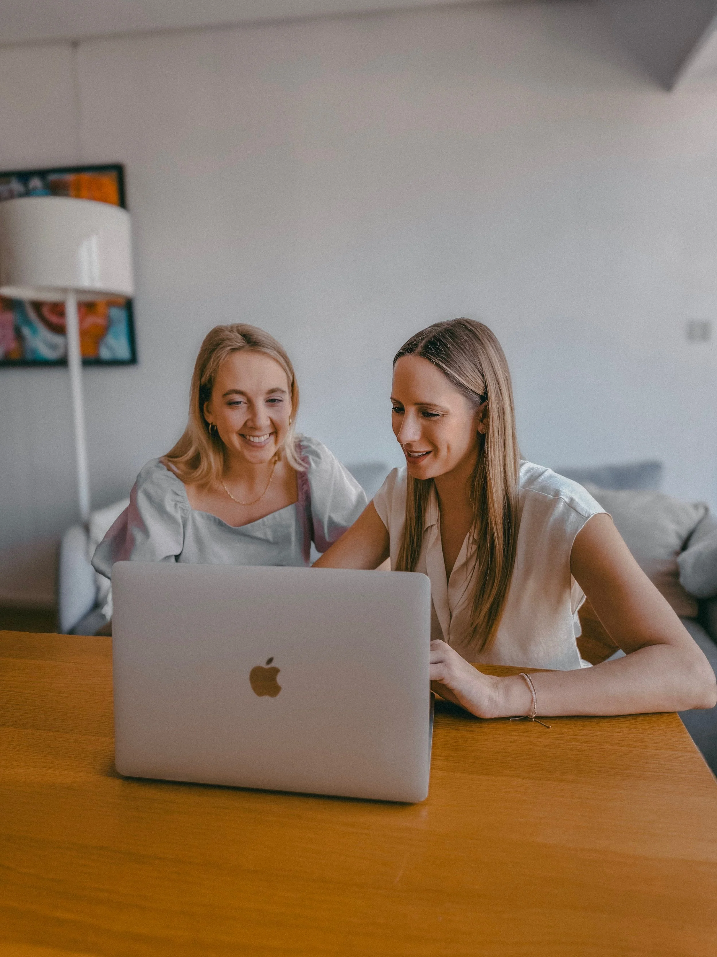 Two wedding and event planners smiling while using a laptop in a living room setting with a sofa and a floor lamp.