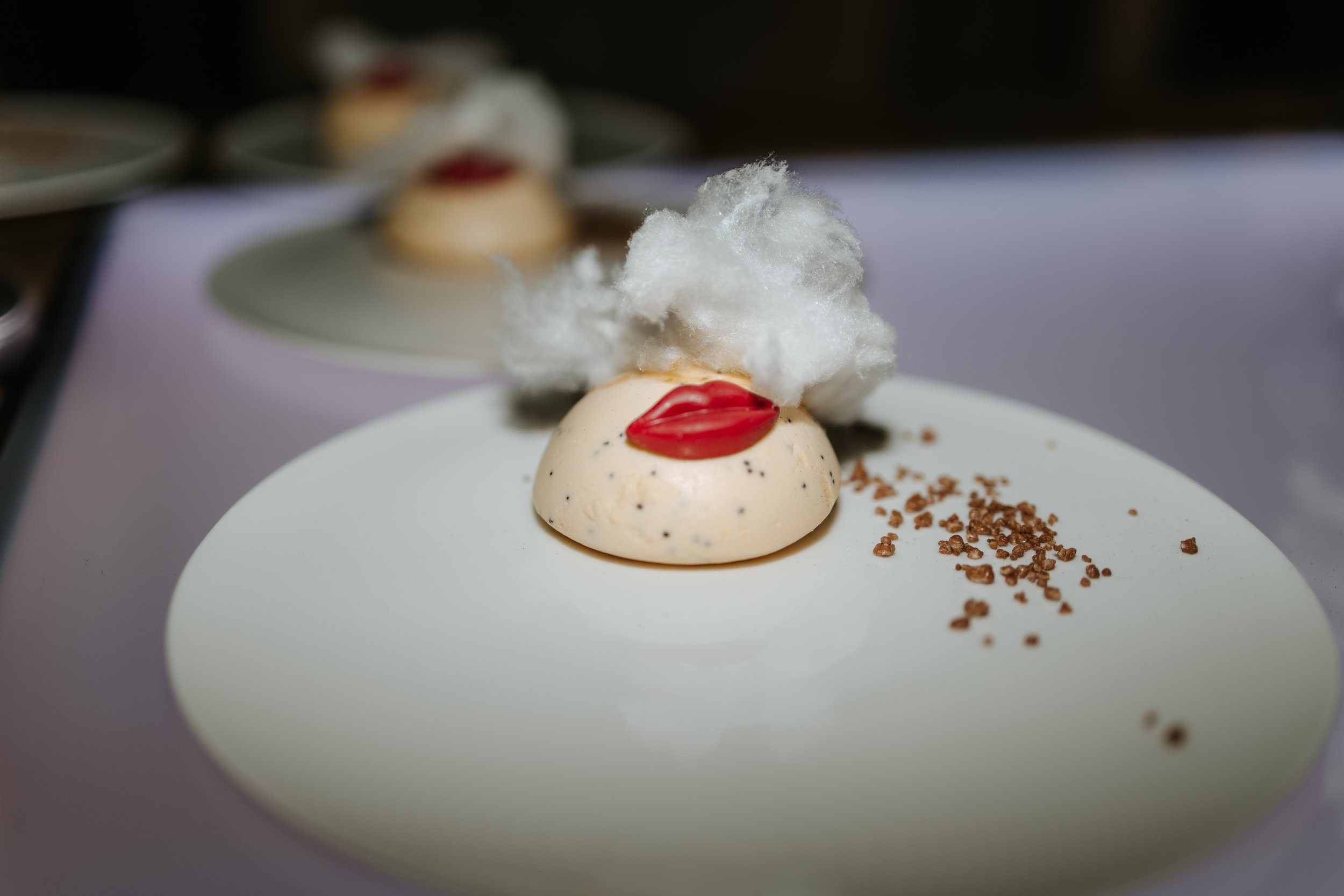 Creative wedding dinner dessert with white mousse, red lips garnish, and cotton candy, served on a white plate with crumbled cookie topping.