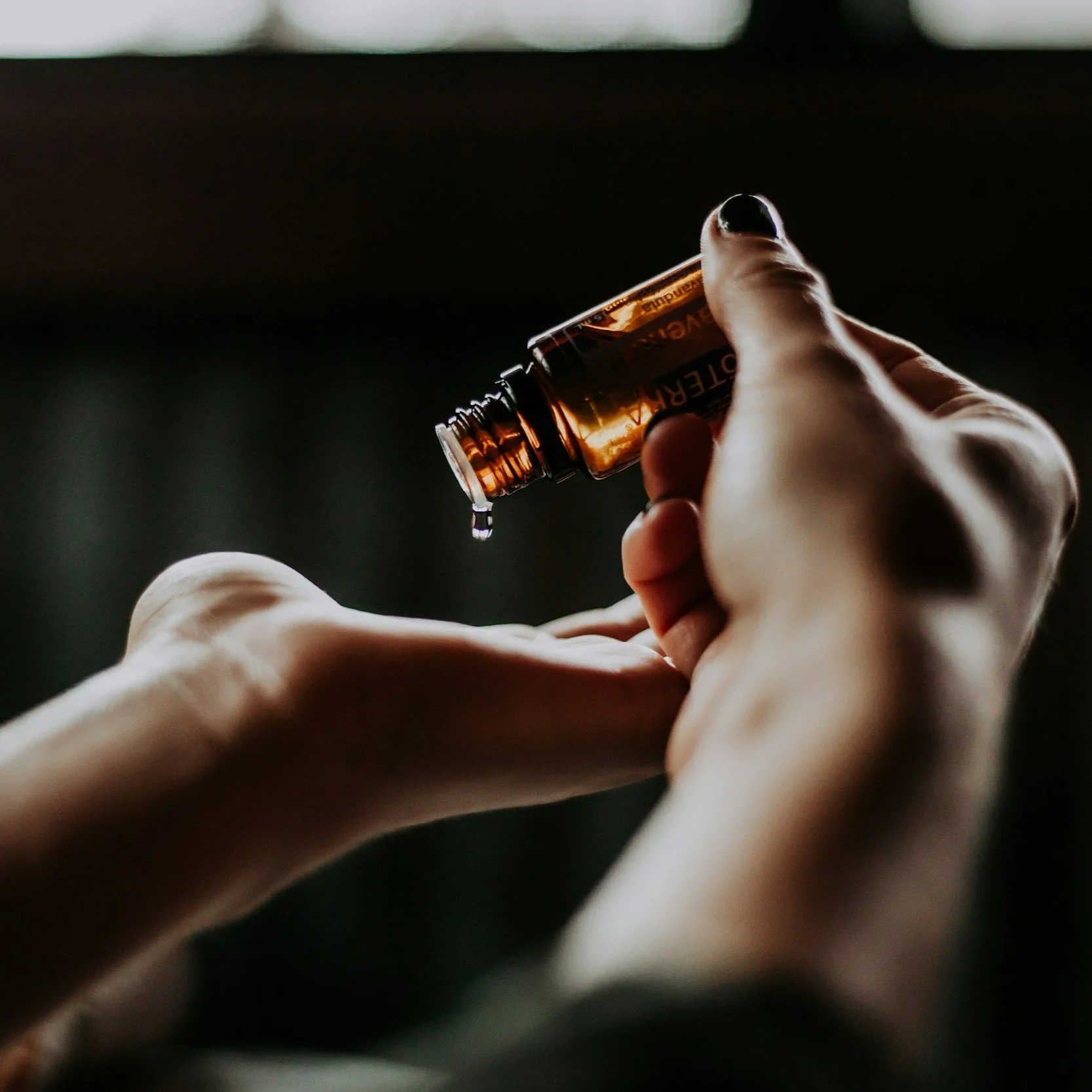 A person's hands holding a small brown glass bottle labeled 'Essential' with a drop of liquid falling from its opening.