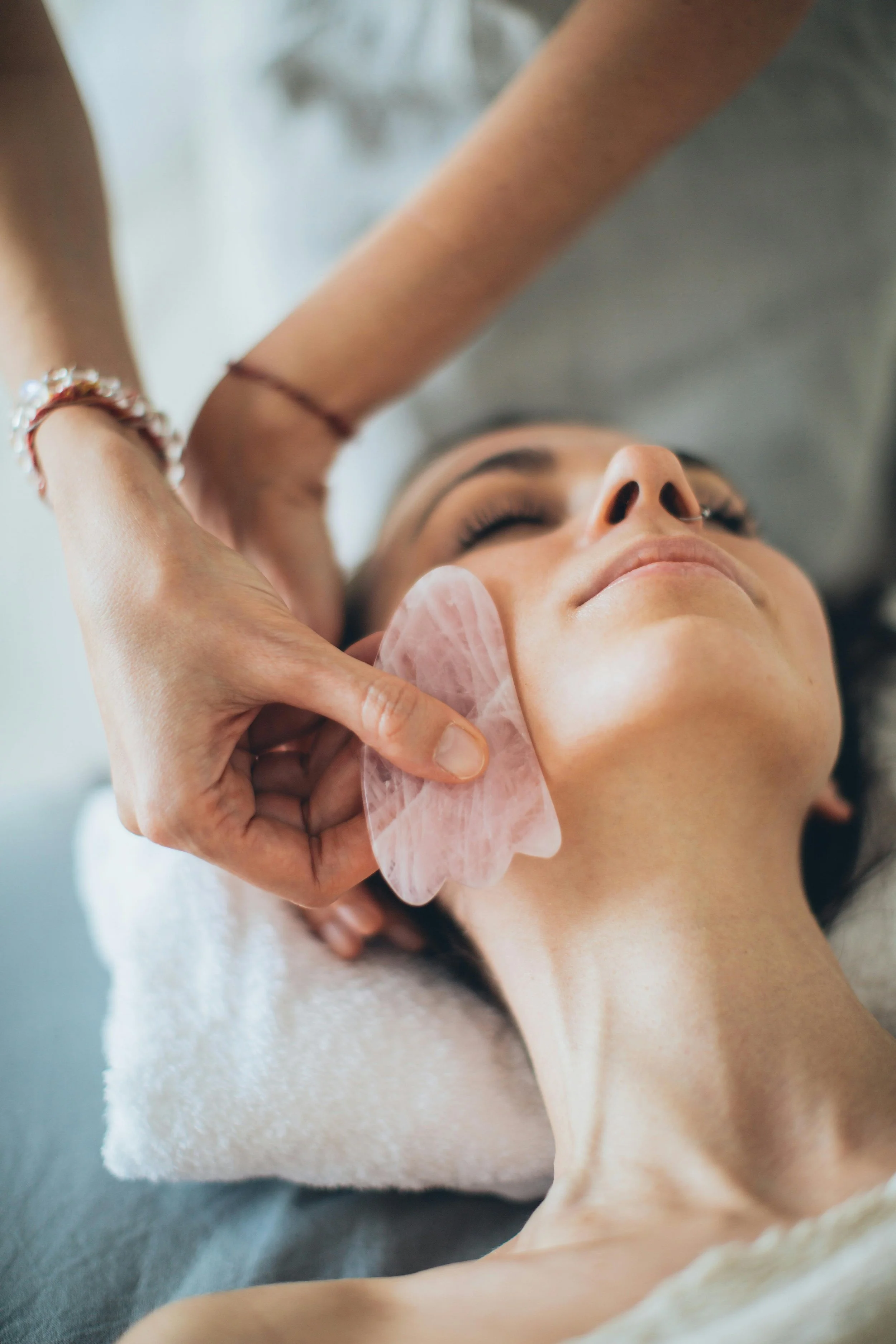 A woman is receiving a facial treatment, with her eyes closed and a pink facial mask applied to her face. An esthetician is gently applying a facial gua sha stone to her cheek.