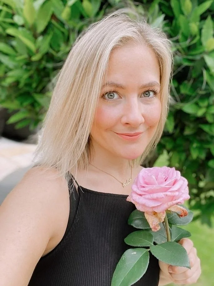 A woman with blonde hair, blue eyes, and a black sleeveless top holding a pink rose outdoors with green foliage in the background.