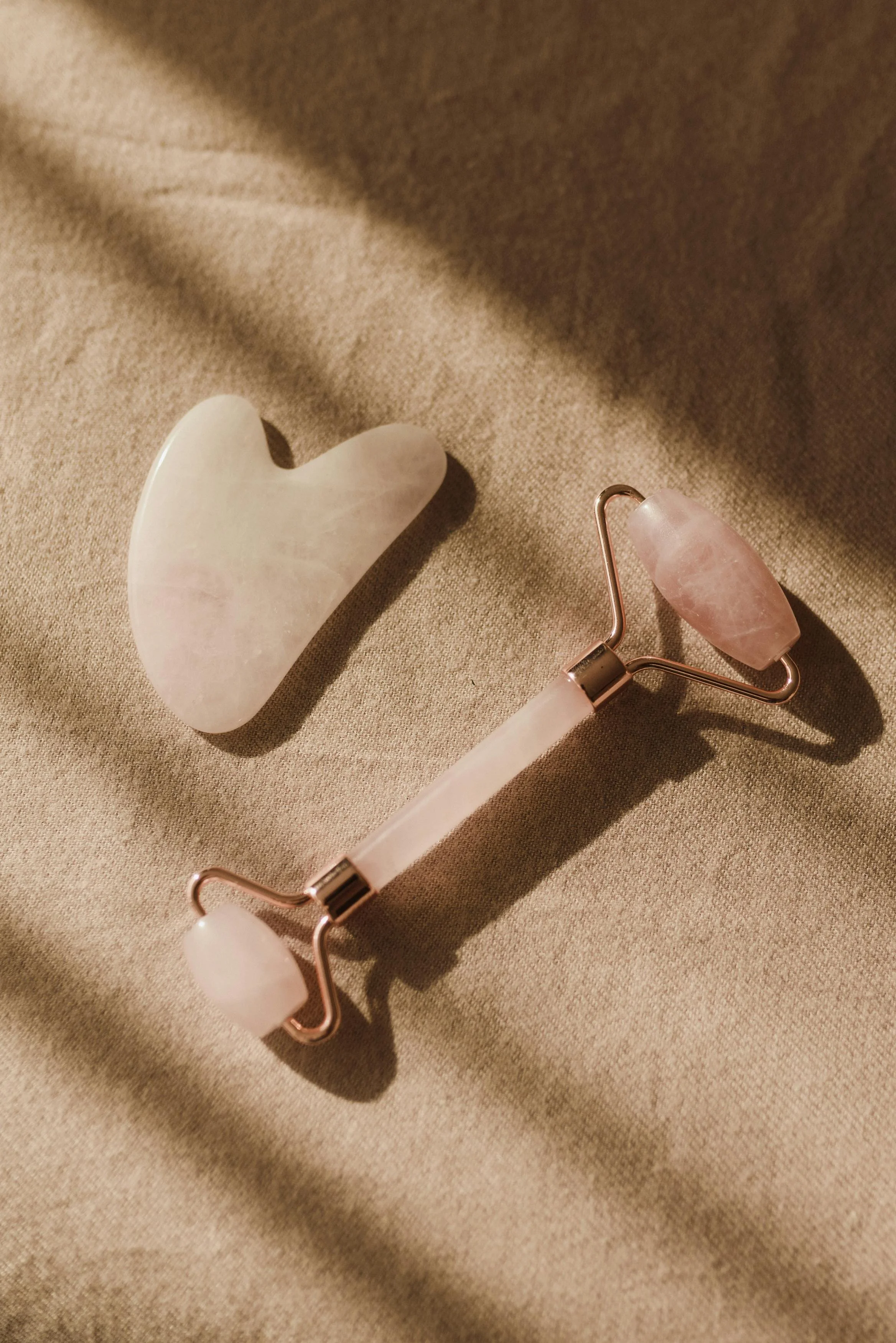 A rose quartz heart-shaped stone and a rose quartz facial roller on a beige fabric surface, casting soft shadows.