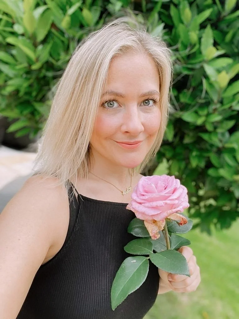 A woman with blonde hair holding a pink rose outdoors in front of green foliage.