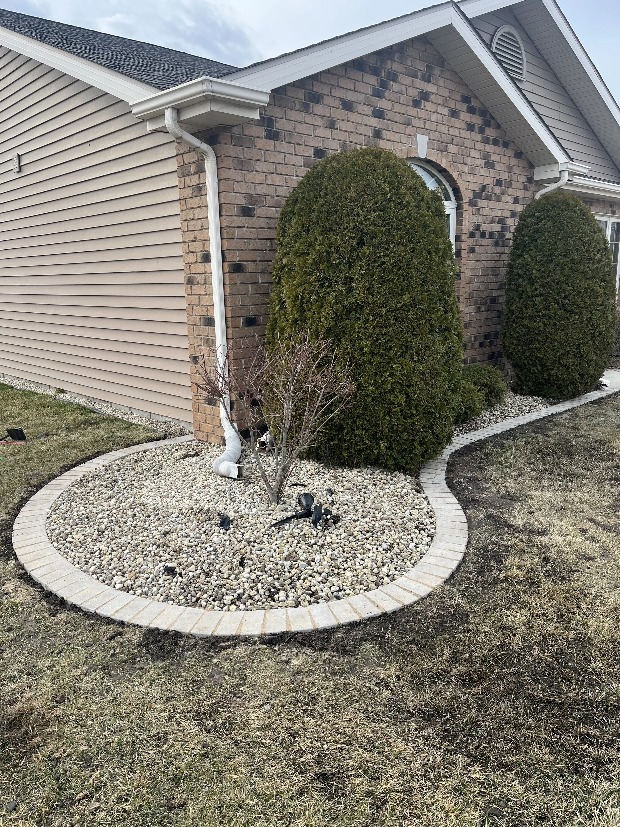 Front yard of a house with a landscaped flower bed surrounded by brick border, containing two green bushes, a small leafless tree, and outdoor lighting, with a house wall and gutter in the background.