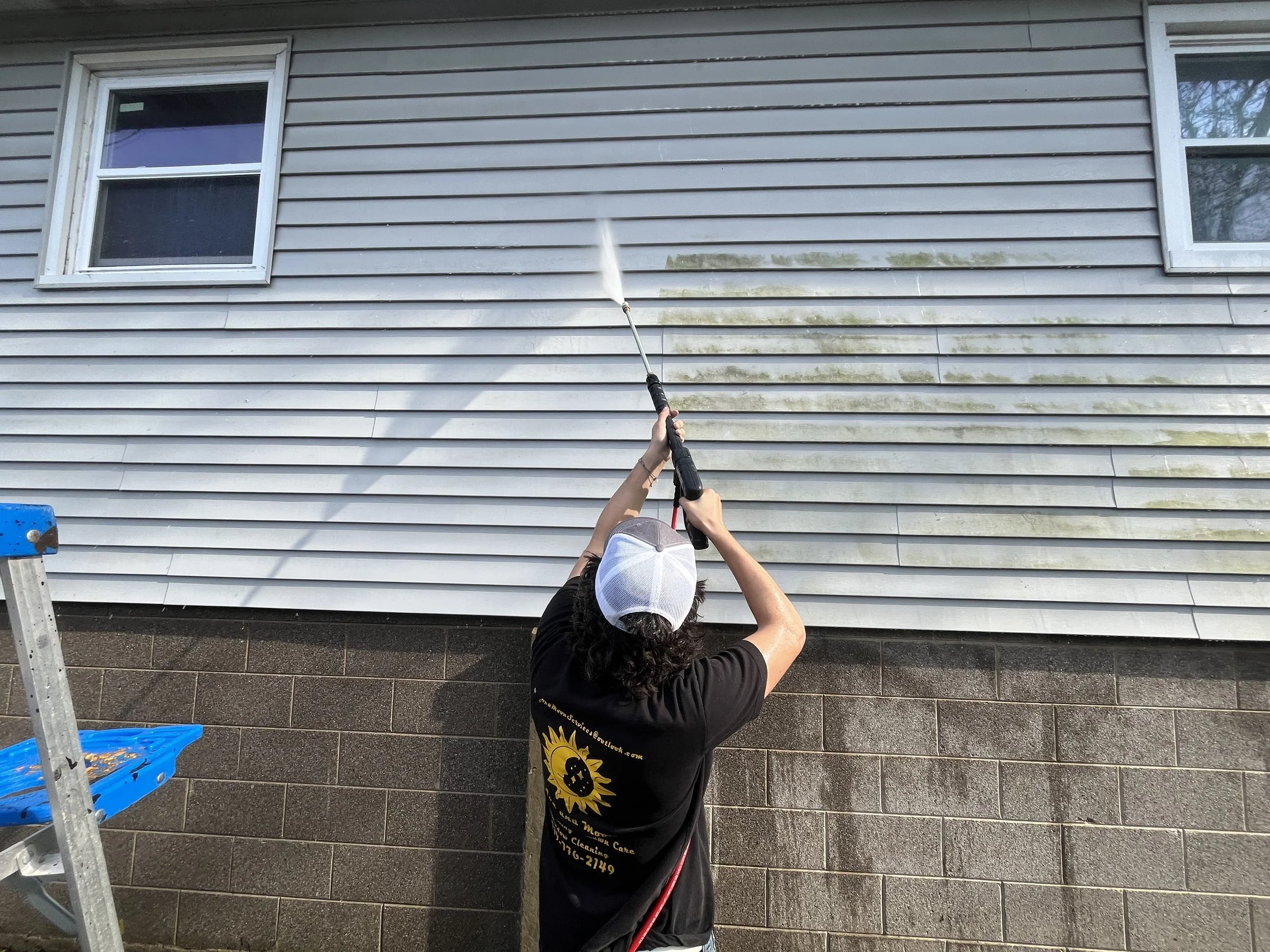 A person pressure washing the exterior wall of a house with horizontal siding, with wet and dirty areas visible.