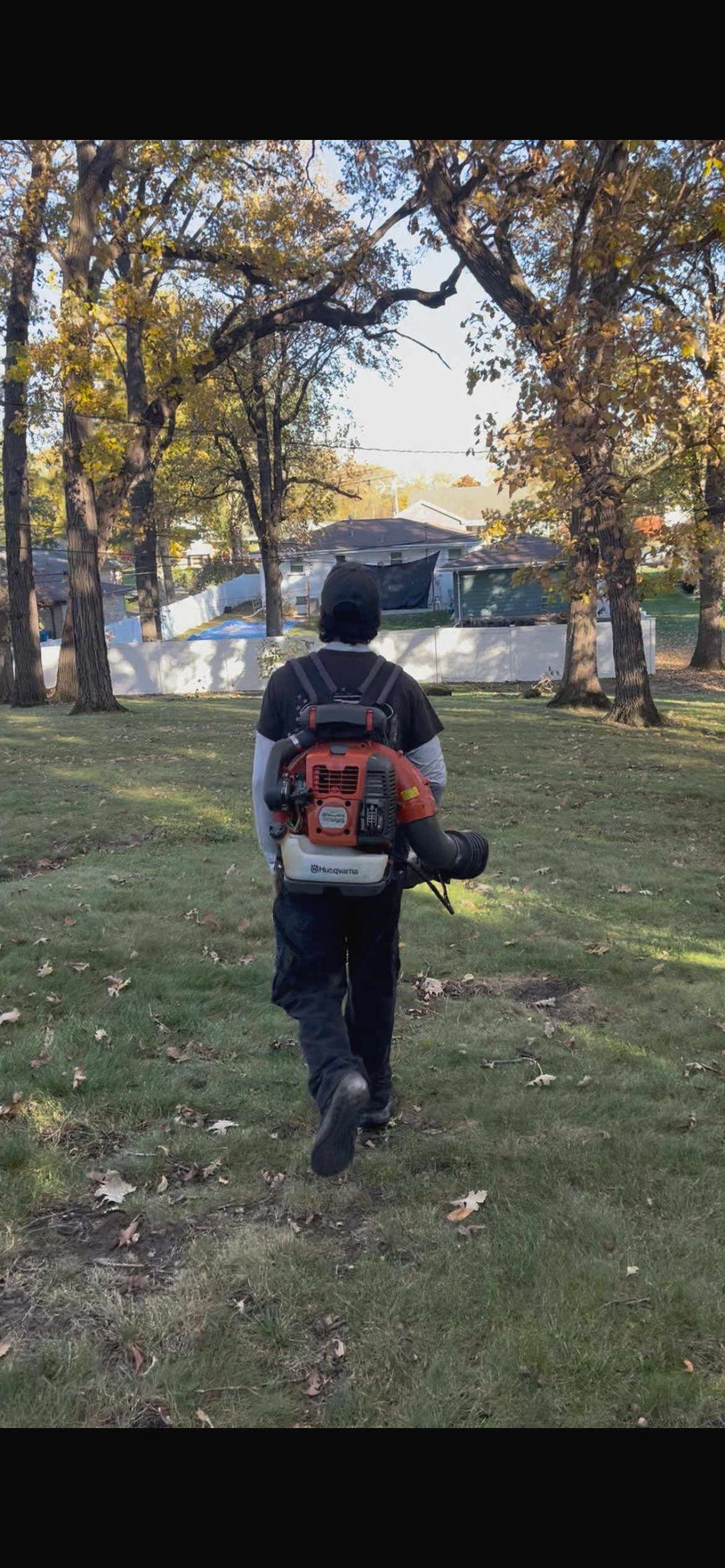Person walking in a yard with trees, carrying a backpack leaf blower.