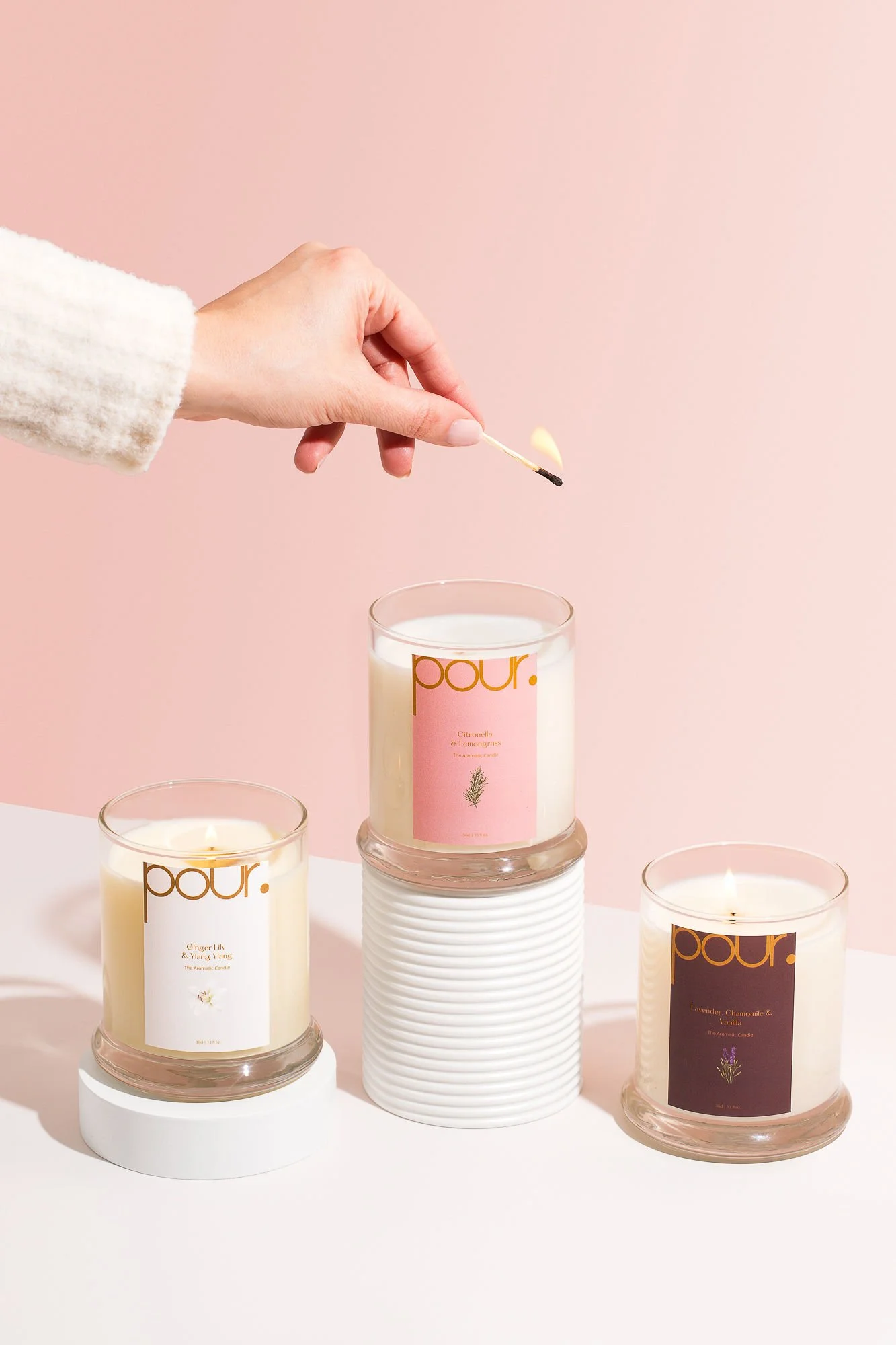 A hand lighting three scented candles with a match on a white table against a pink background.