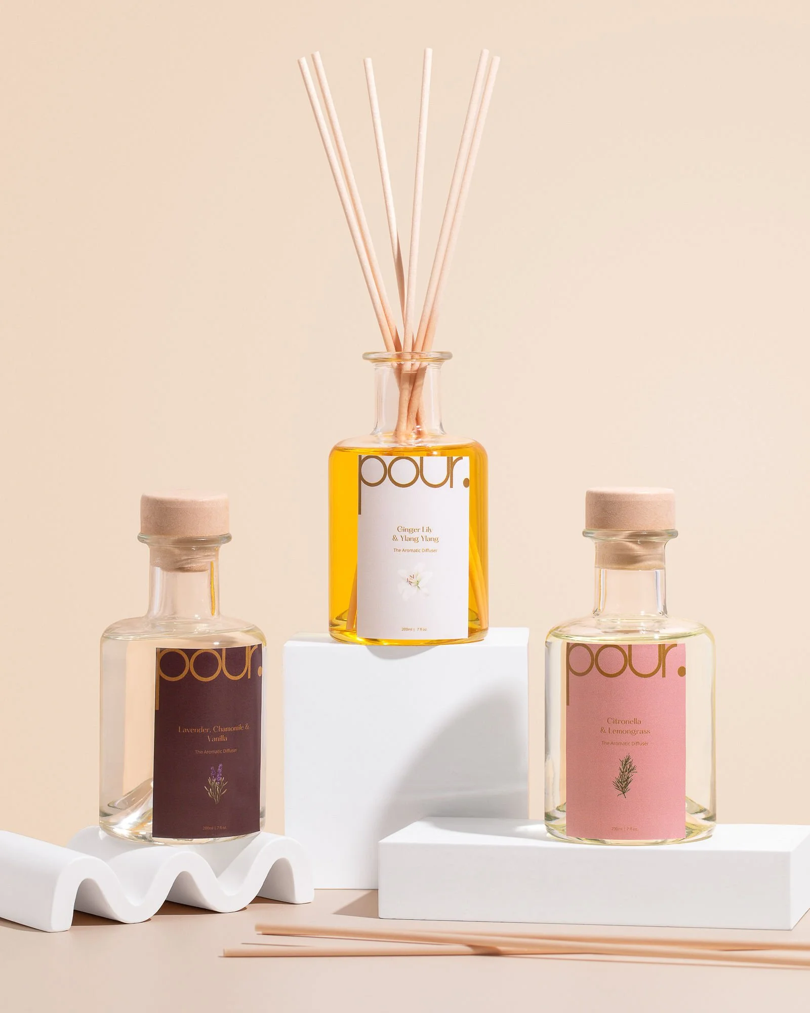 Three aroma diffuser bottles with various scents on a beige background. The central bottle contains ginger lily and ylang ylang, featuring wooden reeds. The left bottle is lavender, chamomile, and vanilla, and the right bottle is citronella and lemongrass. The bottles are creatively arranged on white blocks.