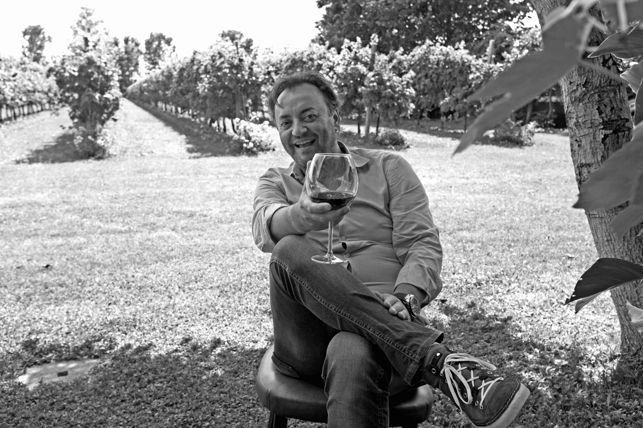 Uomo seduto su una sedia di fronte a un vigneto, che sorseggia un bicchiere di vino rosso, sorridente, in un ambiente esterno con alberi e piante.