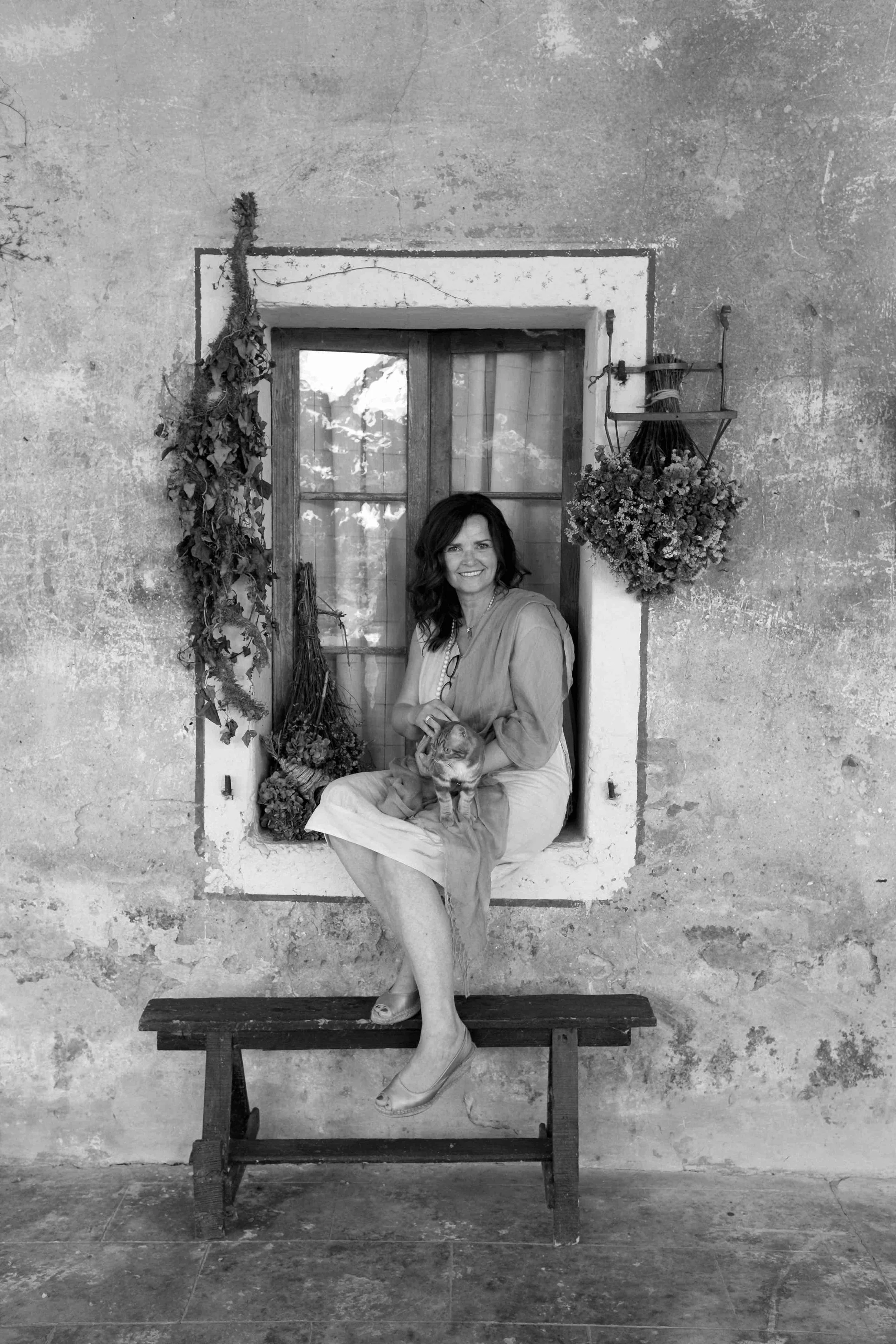 Una donna sorridente seduta su una finestra con un gatto, sullo sfondo un muro antico e decorazioni di fiori secchi.