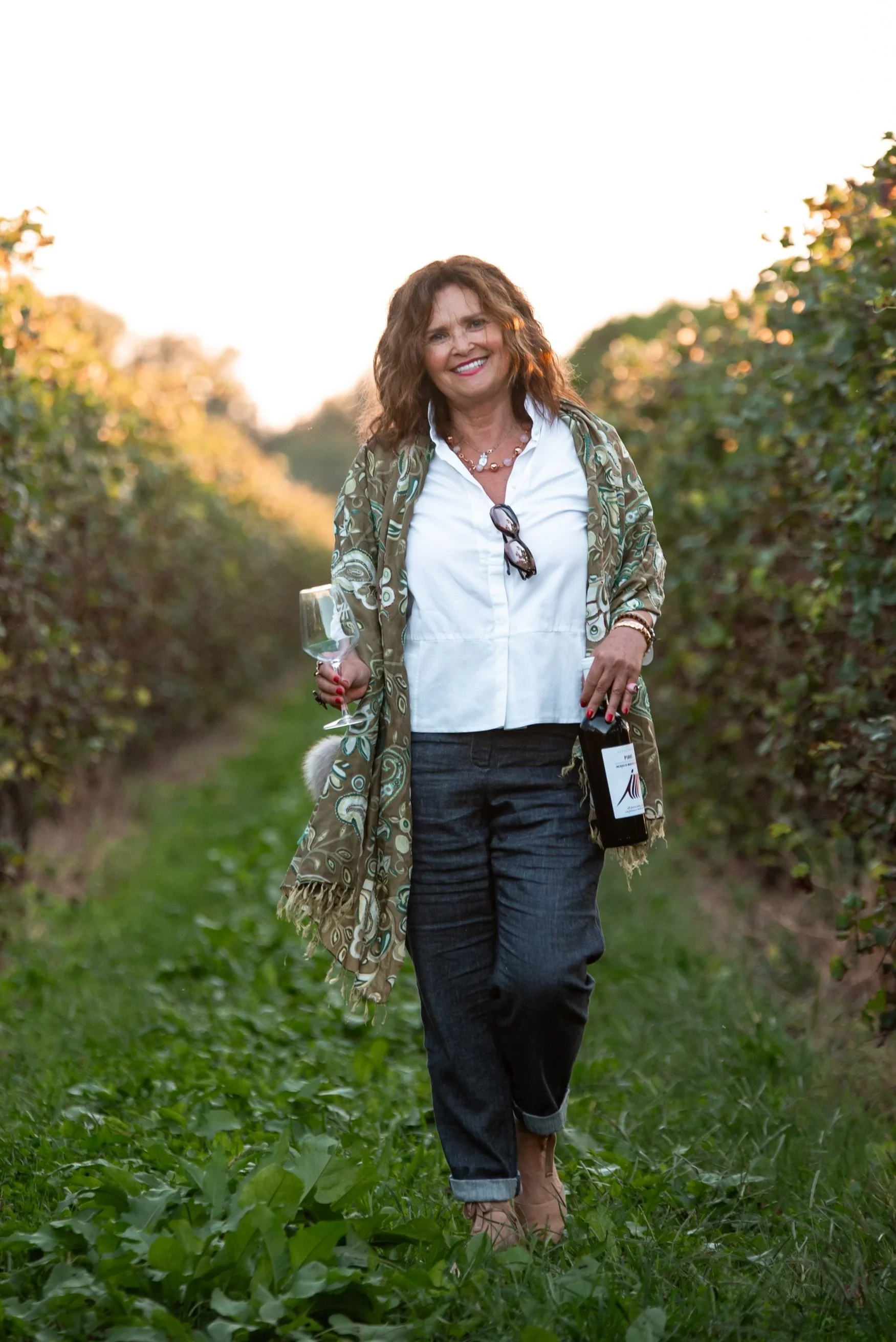 Donna sorridente che cammina tra le viti in un vigneto, tiene un bicchiere di vino e una bottiglia.