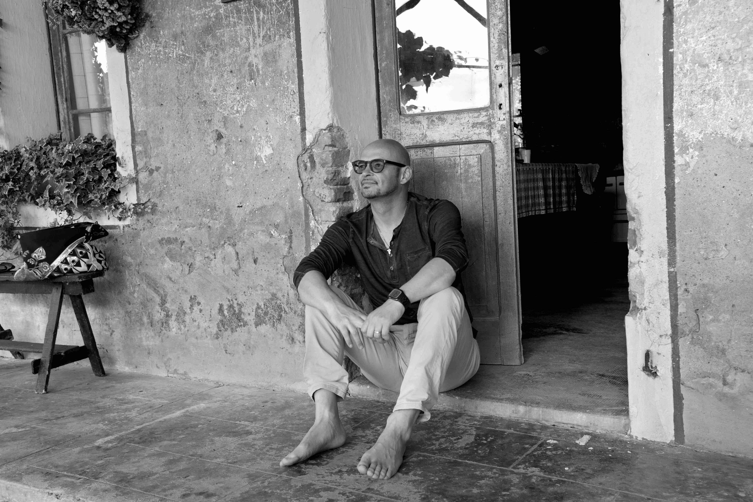 Uomo seduto per terra davanti a una porta in una parete di mattoni e cemento, con pantaloni chiari, camicia nera, occhiali, e senza scarpe. Accanto a una piccola sedia con una borsa sopra.