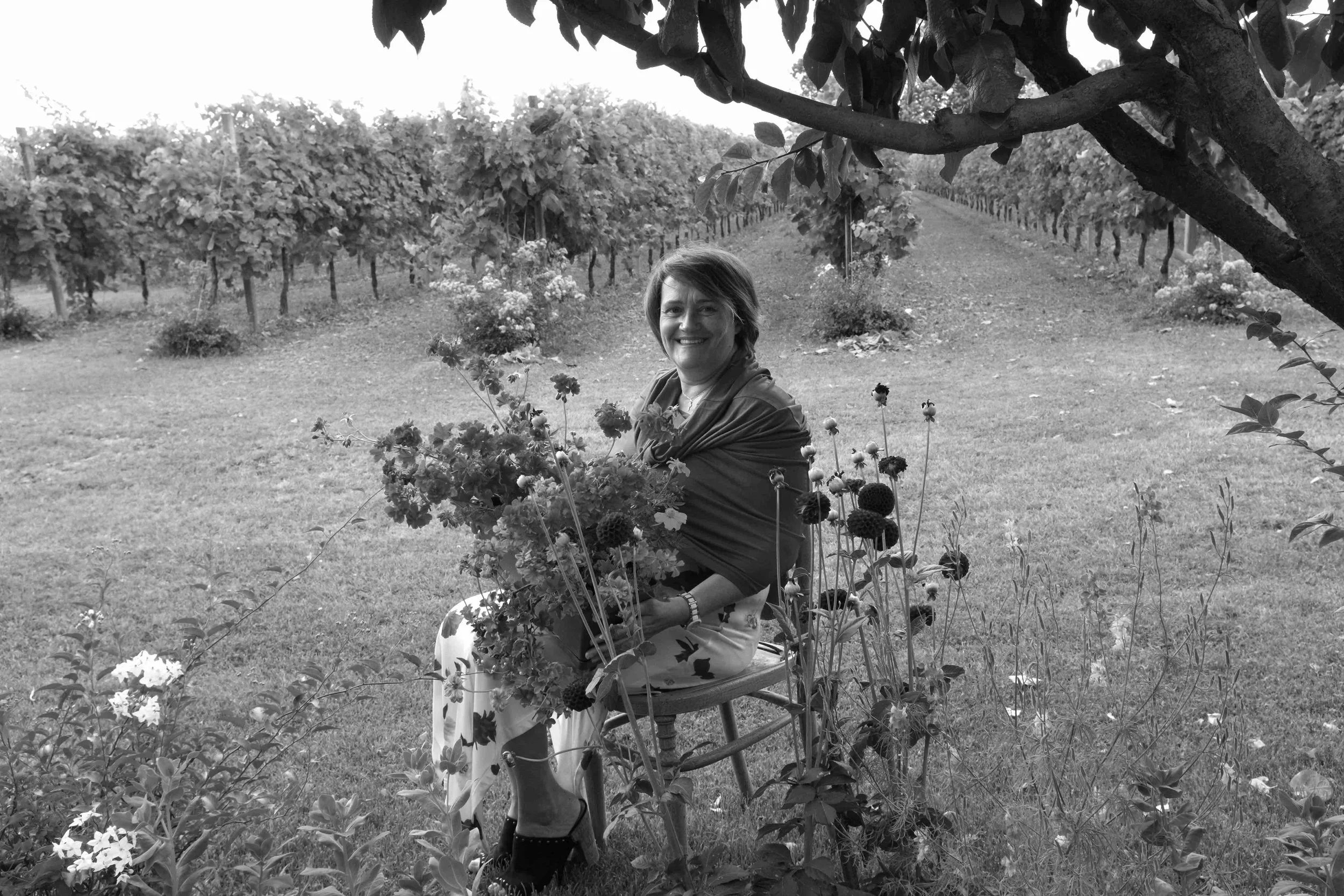 Donna seduta su una sedia in un campo di fiori e viti, circondata da piante e alberi, sorridente con uno sfondo di viti in file.