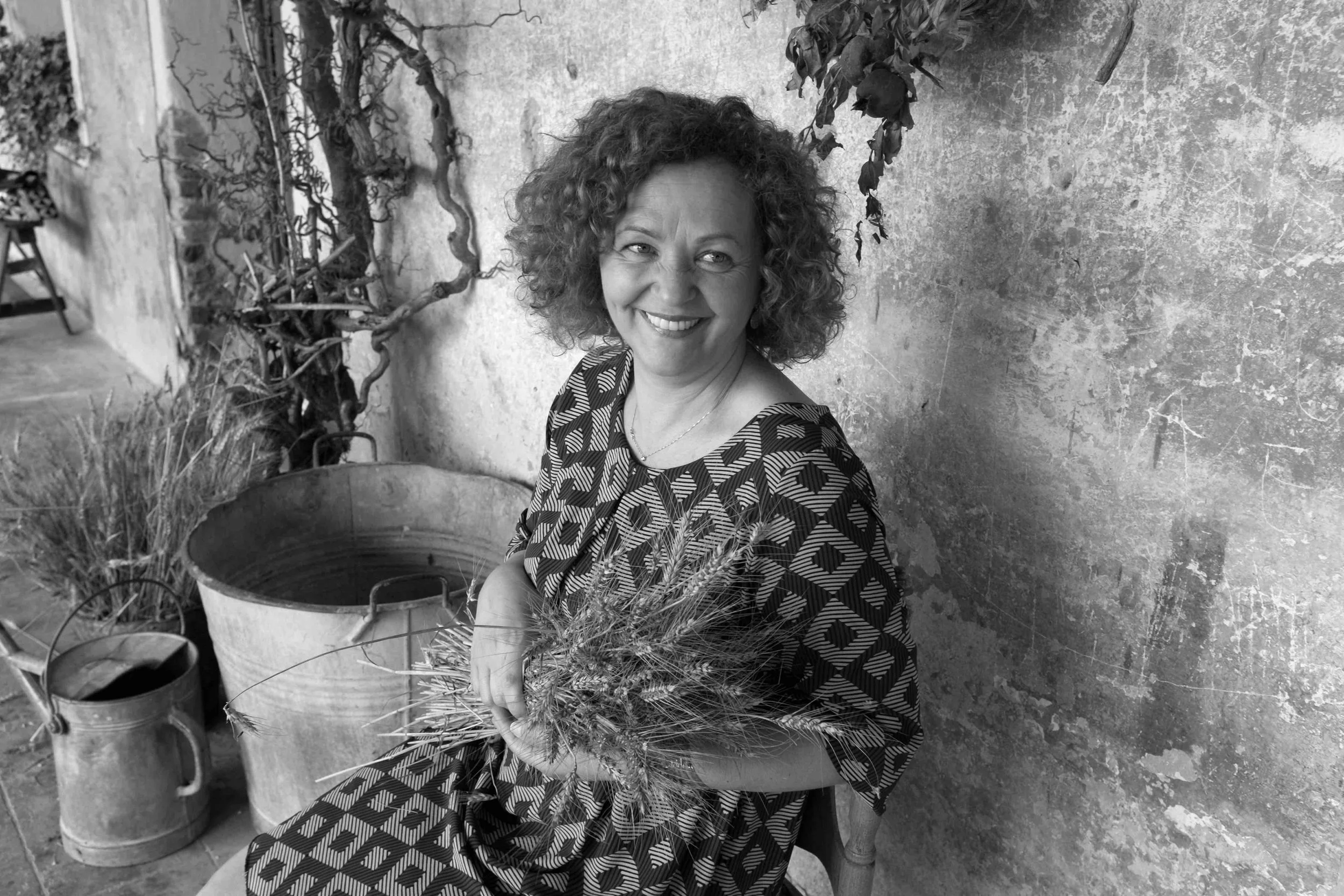 Una donna sorridente seduta vicino a un muro con piante, tiene un mazzo di spighe di grano, l'immagine è in bianco e nero.