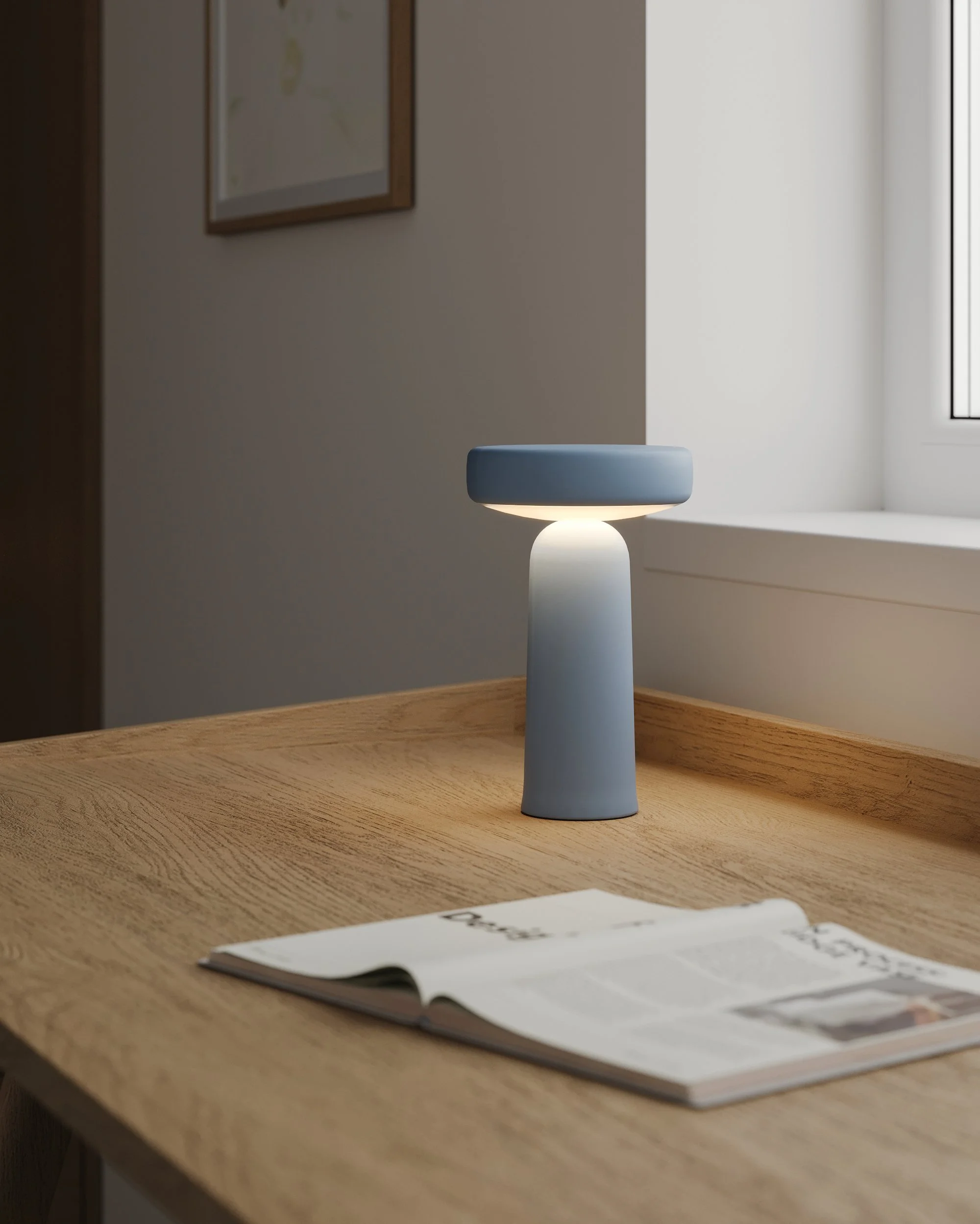 Residential interior design. Minimalist blue table lamp on a light oak desk beside a window in a calm Scandinavian-style interior.