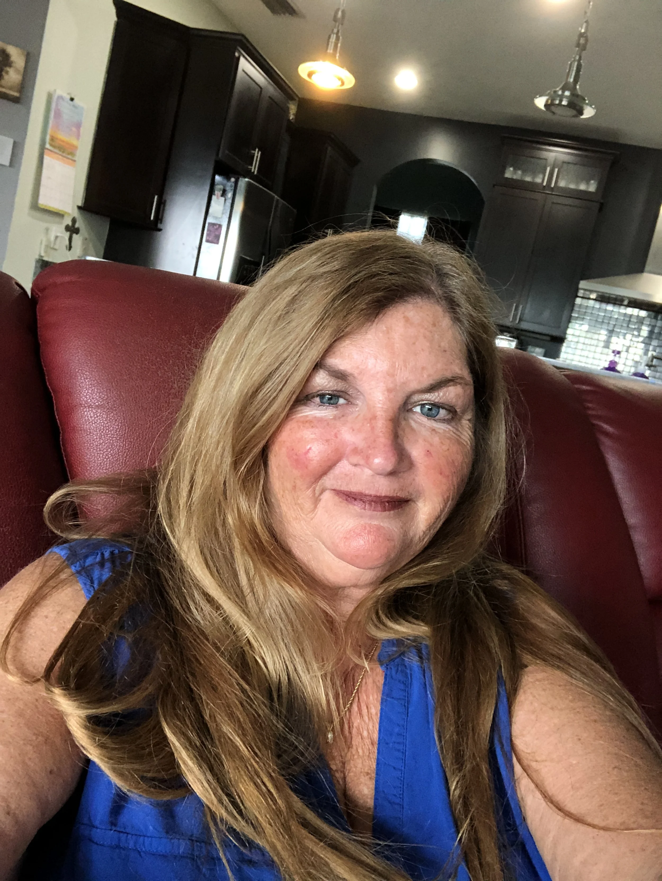 A woman with long blonde hair and blue eyes taking a selfie inside a house. She is sitting on a red leather couch, wearing a blue top, with a kitchen in the background featuring dark cabinets and stainless steel appliances.