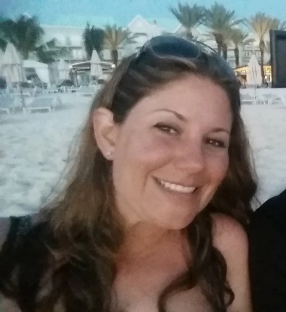 Smiling woman with wavy brown hair, wearing sunglasses on her head, at the beach with palm trees, lounge chairs, and a white building in the background.