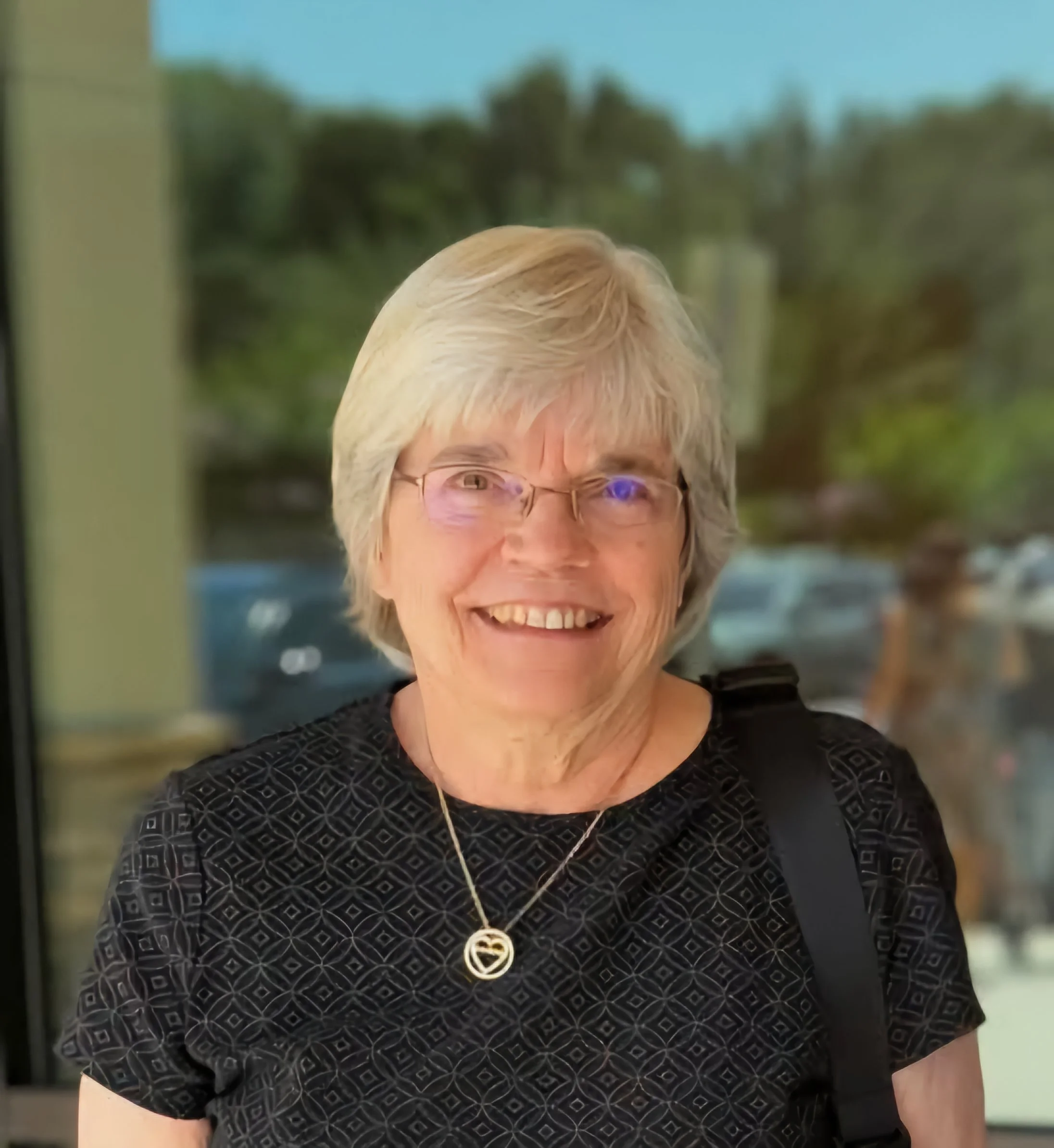 An elderly woman with short white hair, wearing glasses, a black patterned shirt, a necklace, and a shoulder bag, smiling in front of a blurred outdoor background.