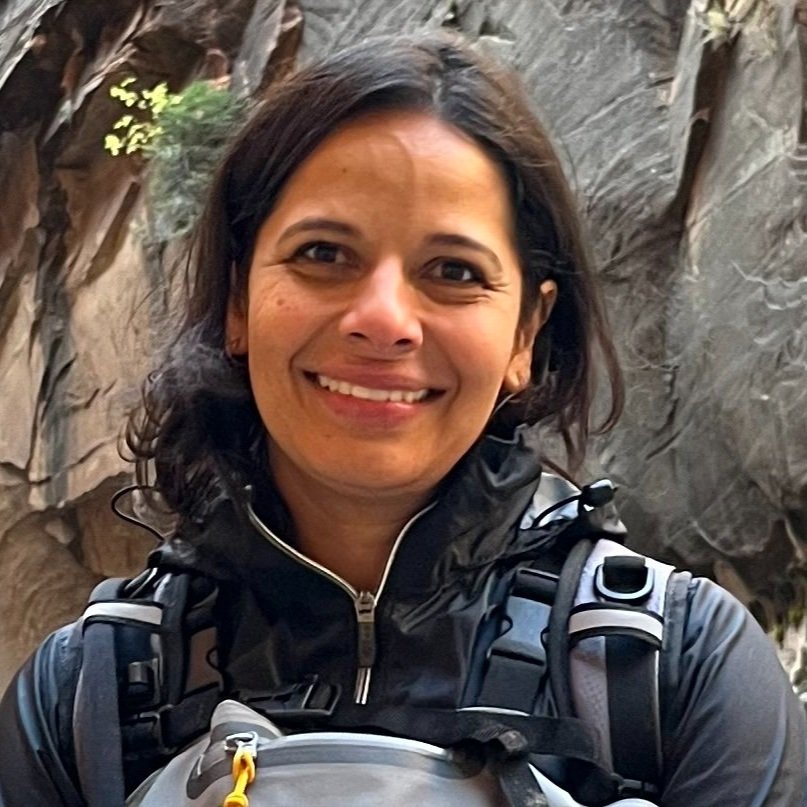 A woman with dark hair smiling outdoors in a rocky canyon or gorge, wearing a black jacket and a backpack.