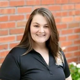 Young woman with long brown hair smiling in front of a brick wall wearing a black polo shirt.