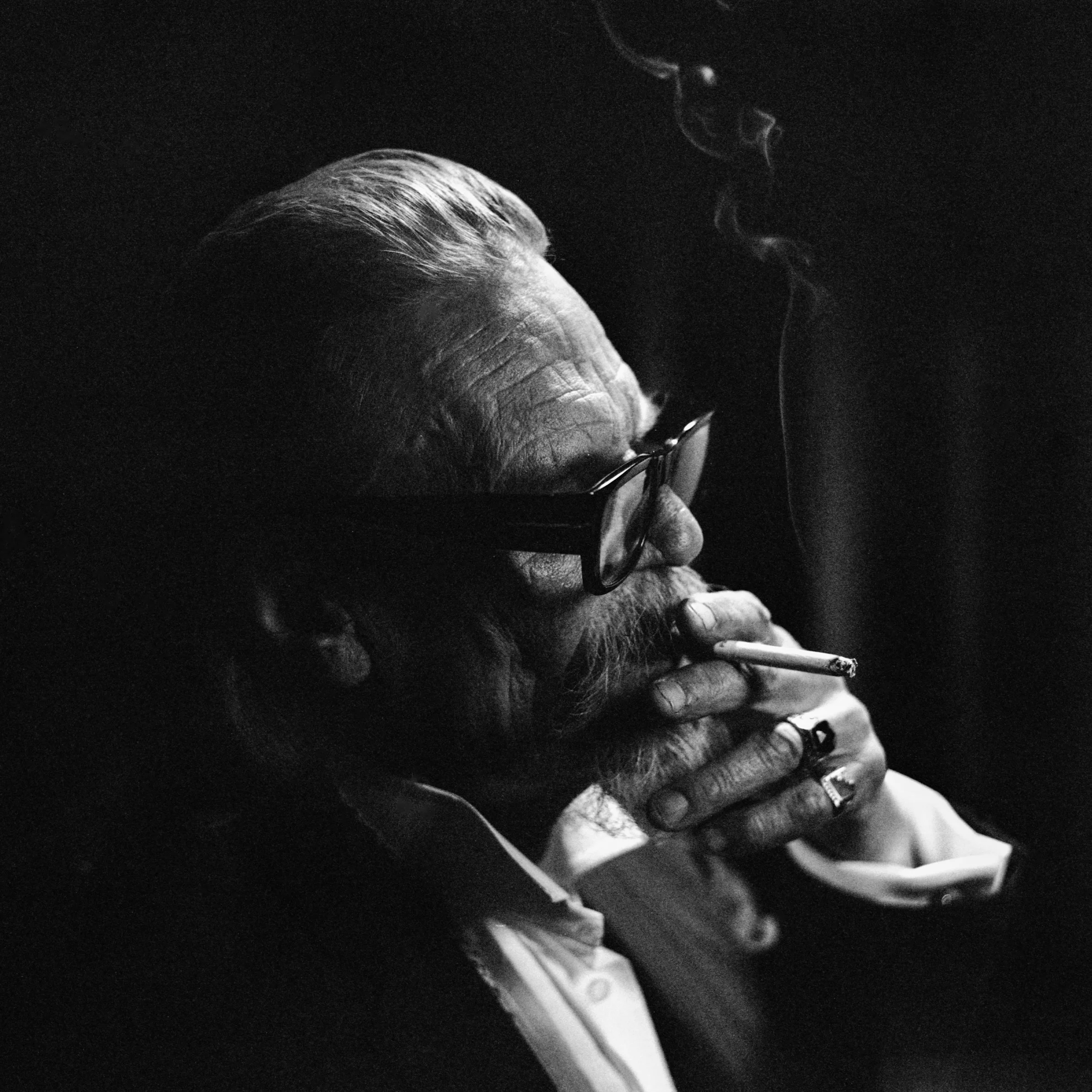 A black and white photo of an elderly man with glasses smoking a cigarette.