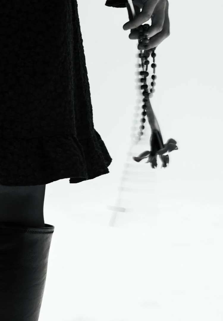 A hand holding a rosary with a cross, visible from a low angle, against a bright background. The lower part of a person wearing a dark coat and tall boots is also visible.