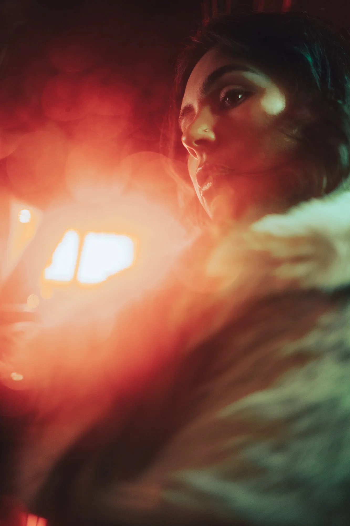 A woman with dark hair and a nose piercing gazes to the right, illuminated by warm, colorful lights, with a blurred background and a bokeh effect.