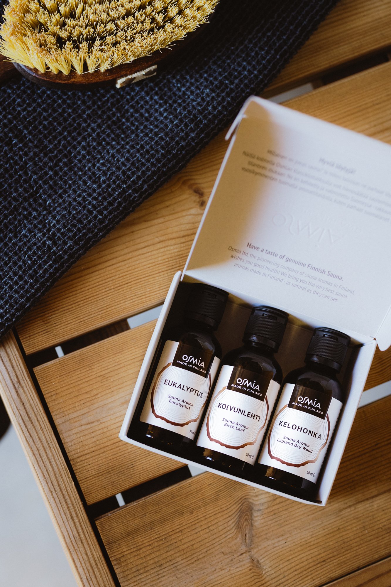 A box containing three bottles of sauna aroma oils labeled eucalyptus, birch leaf, and dry wood, resting on a wooden surface with a dark cloth underneath and a brush visible in the background.