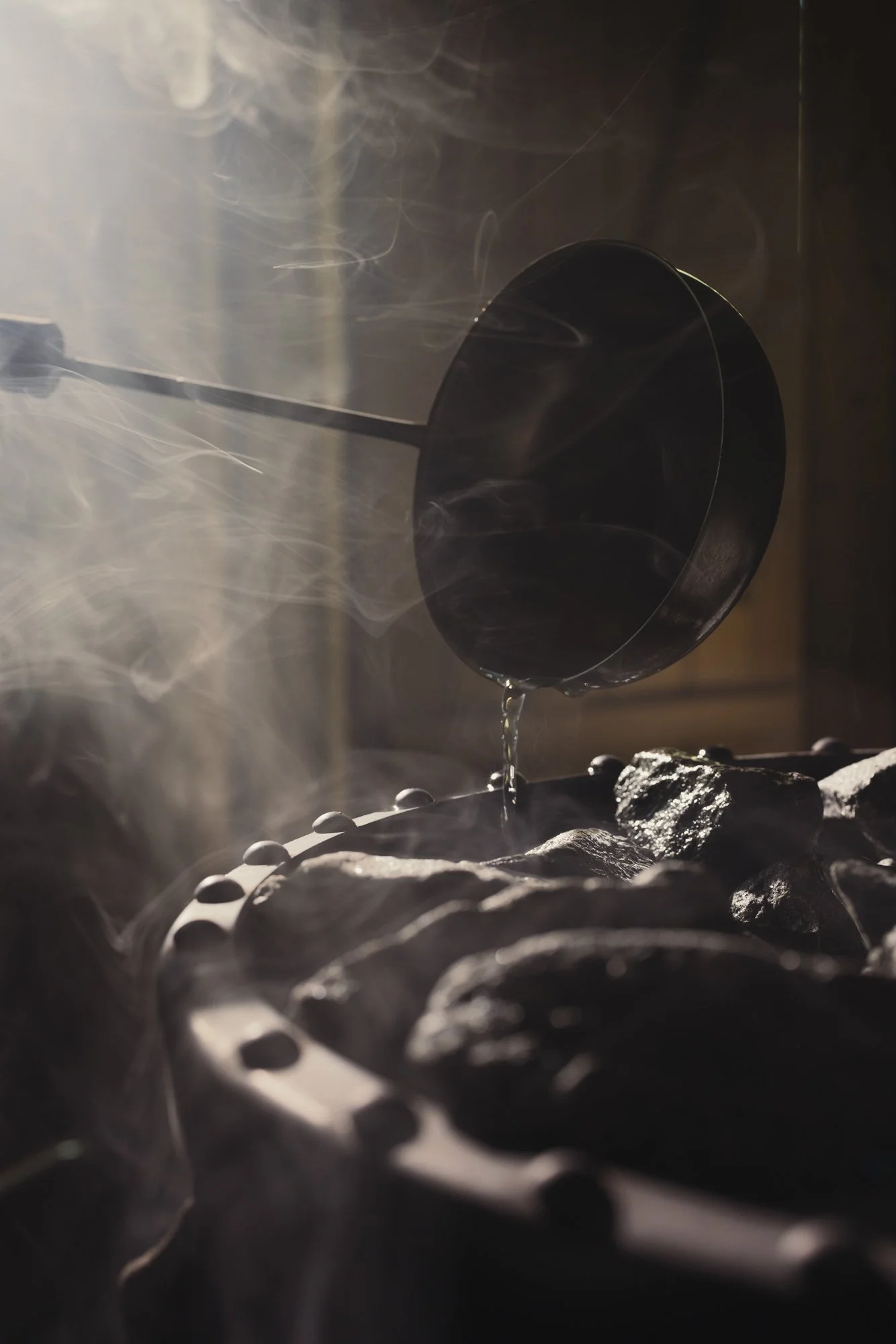 A black ladle hanging above a grill with rocks, with water dripping from it and steam rising around.